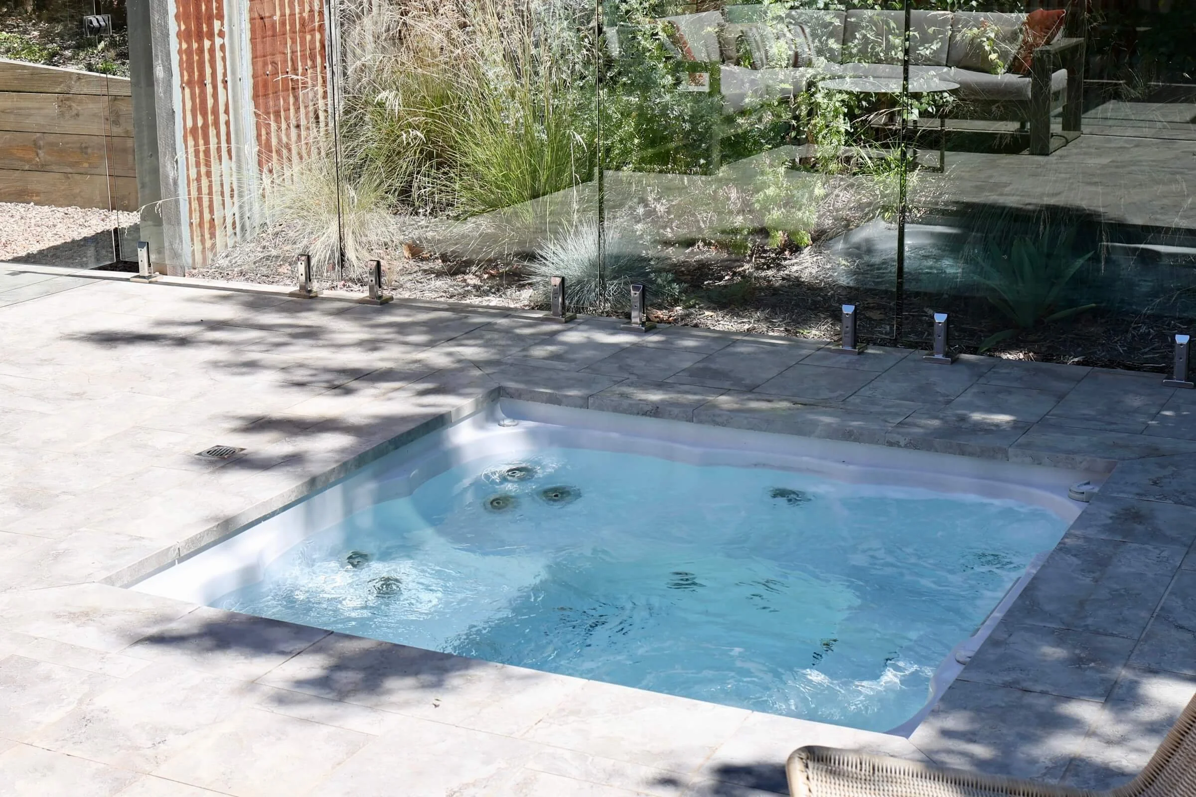A backyard with a rectangular pool with bubbling water, surrounded by light-colored stone paving, enclosed by a glass fence with metal supports, with various plants and a seating area visible in the background.
