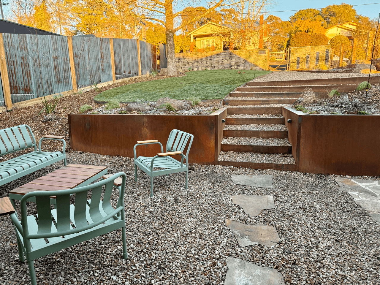 Backyard with gravel ground, metal patio furniture including a bench, two chairs, and a small table, stone pathway, and a set of stairs leading up to a grassy area with a tree, fencing, and residential houses in the background.