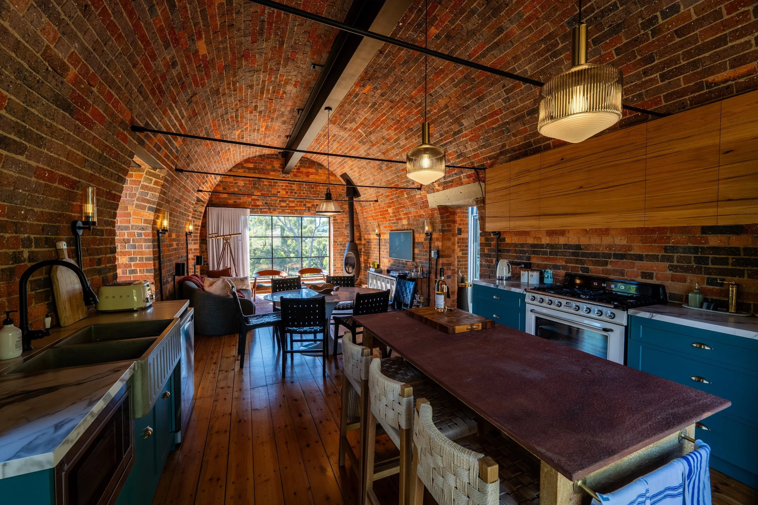 Open-concept brick loft with kitchen and living area, large window, wooden furniture, hanging pendant lights, and brick vaulted ceiling.
