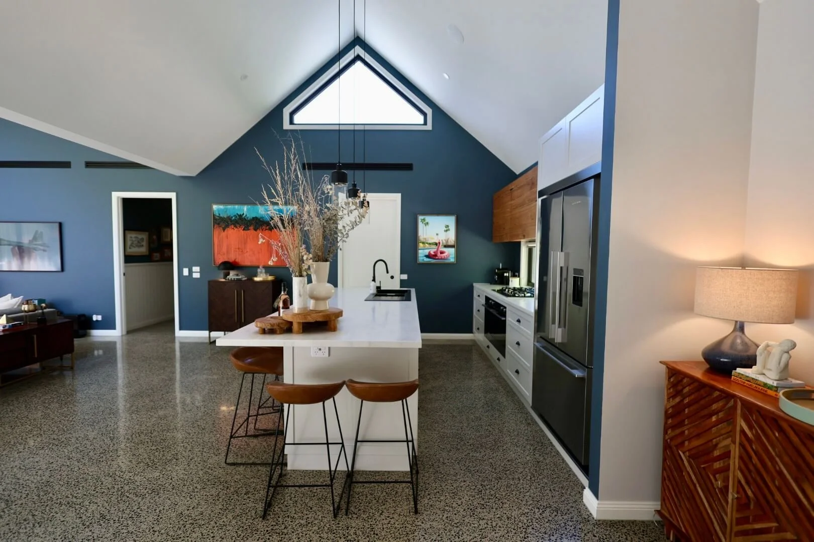 Open-concept kitchen with a white island, blue accent walls, gray cabinets, stainless steel appliances, and decorative elements like vases and artwork. Includes a dining area and living space visible in the background.