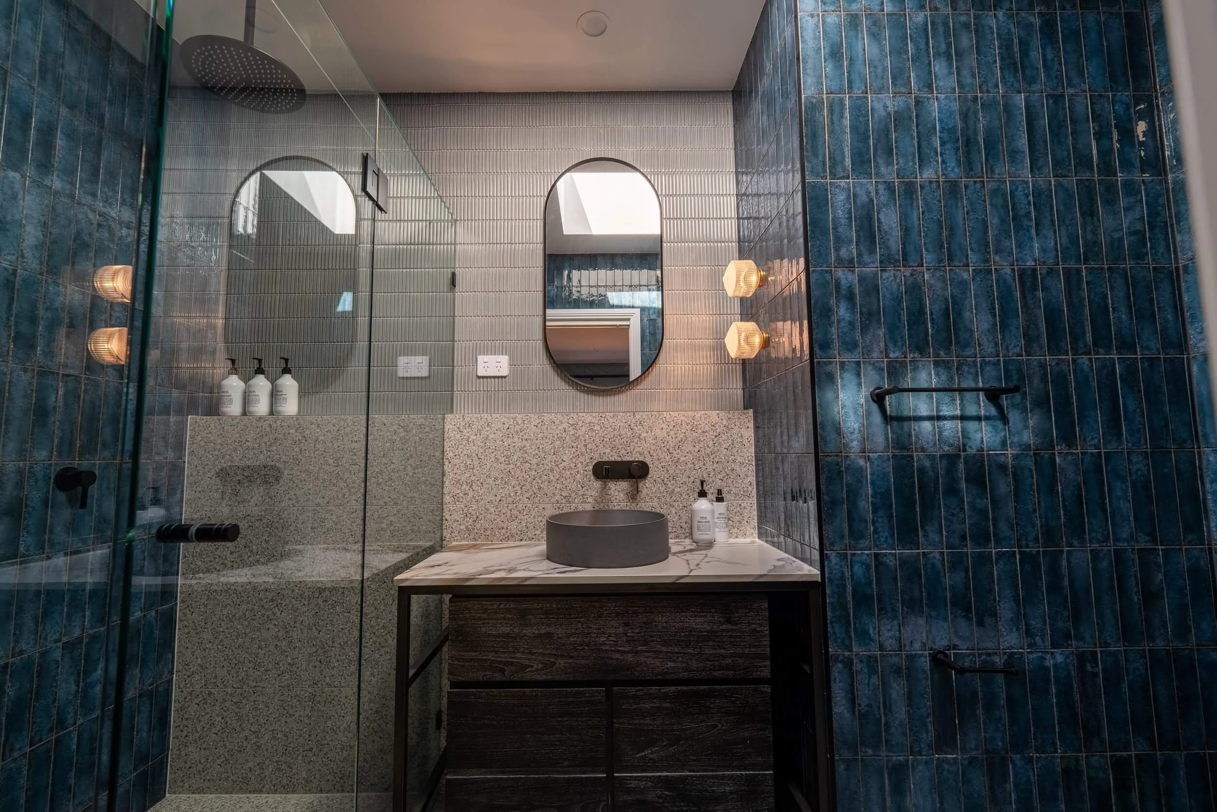 A modern bathroom with blue tiled walls, a marble countertop with a round vessel sink, and a large mirror. There are two bottles of soap on the counter, single light fixtures, and a shower area with dark fixtures.