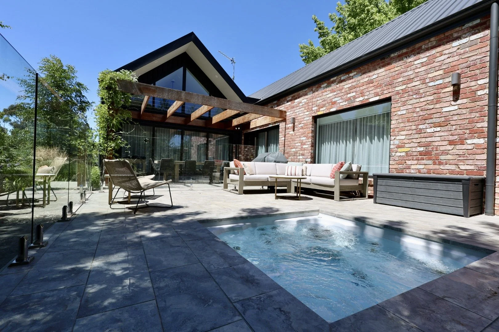 Outdoor patio with small hot tub, lounge chairs, and couches, enclosed by glass fencing, attached to a brick house with large windows and covered patio area, surrounded by trees.