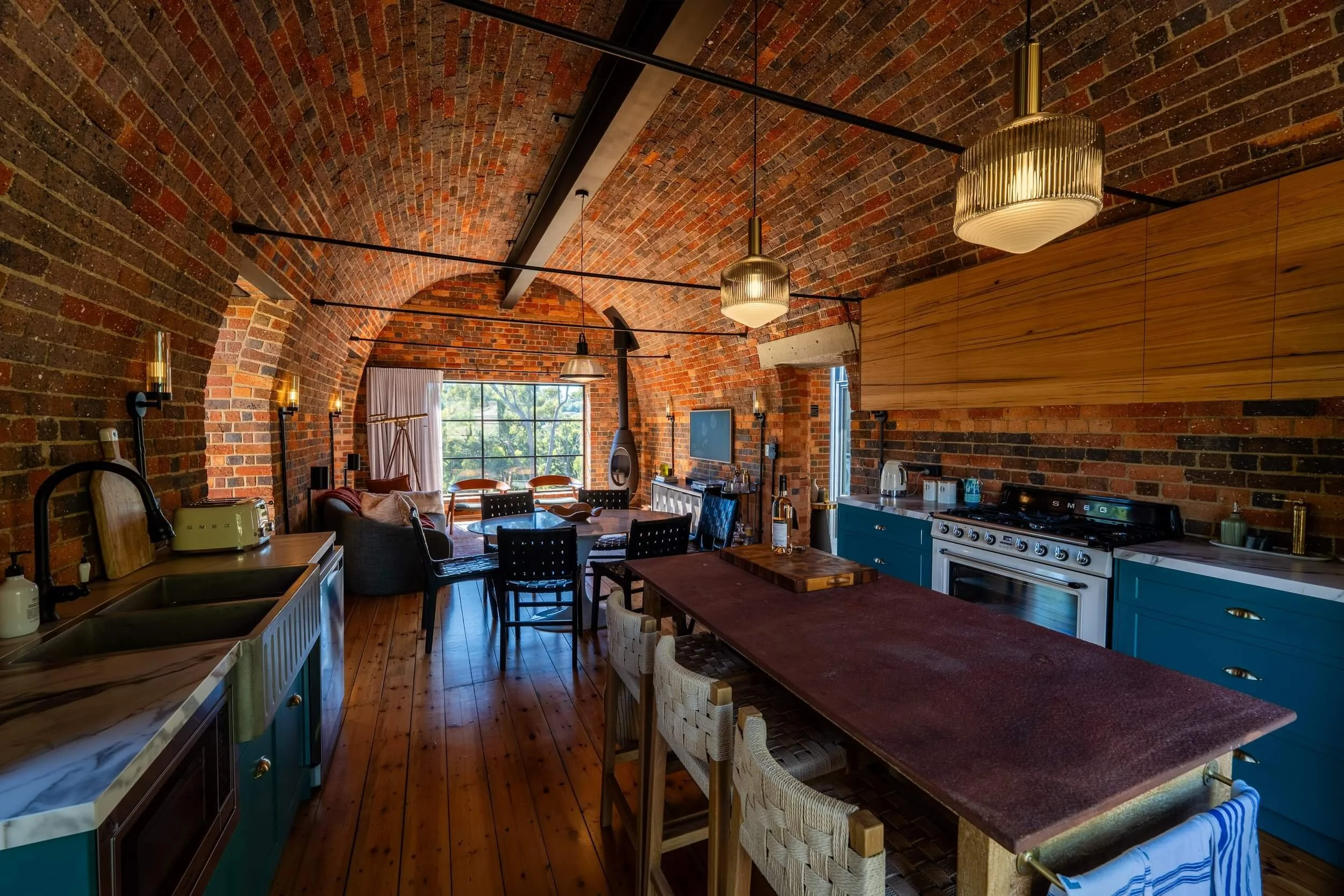 Open-concept kitchen and living space with brick arched ceiling, pendant lights, and large window showing greenery outside. Kitchen features teal cabinets, white marble countertop, and stainless steel appliances. Dining table with woven chairs is in 