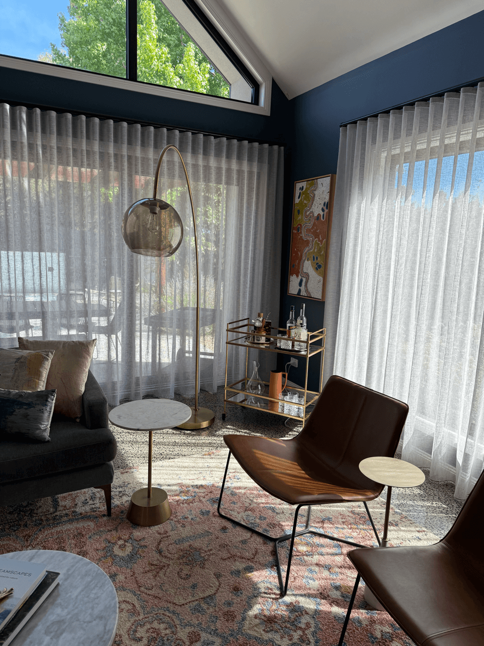 Living room corner with large windows and sheer curtains, a modern floor lamp, a sofa with pillows, two modern chairs with small side tables, a bar cart with bottles and glassware, an abstract art piece on the wall, and a patterned area rug.
