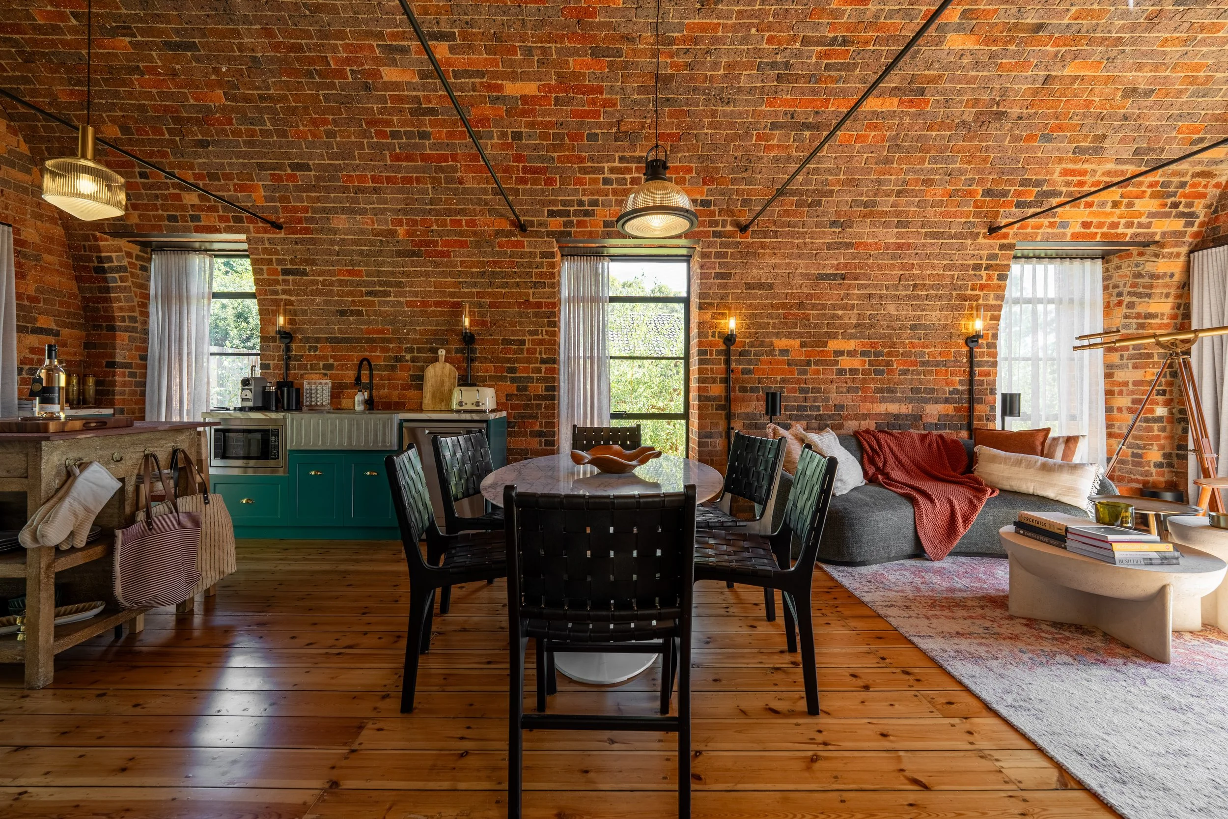 Interior of a rustic living space with exposed brick walls, wooden floors, a dining table with six black chairs, a gray sofa with red and beige cushions, and various decor and furniture pieces.
