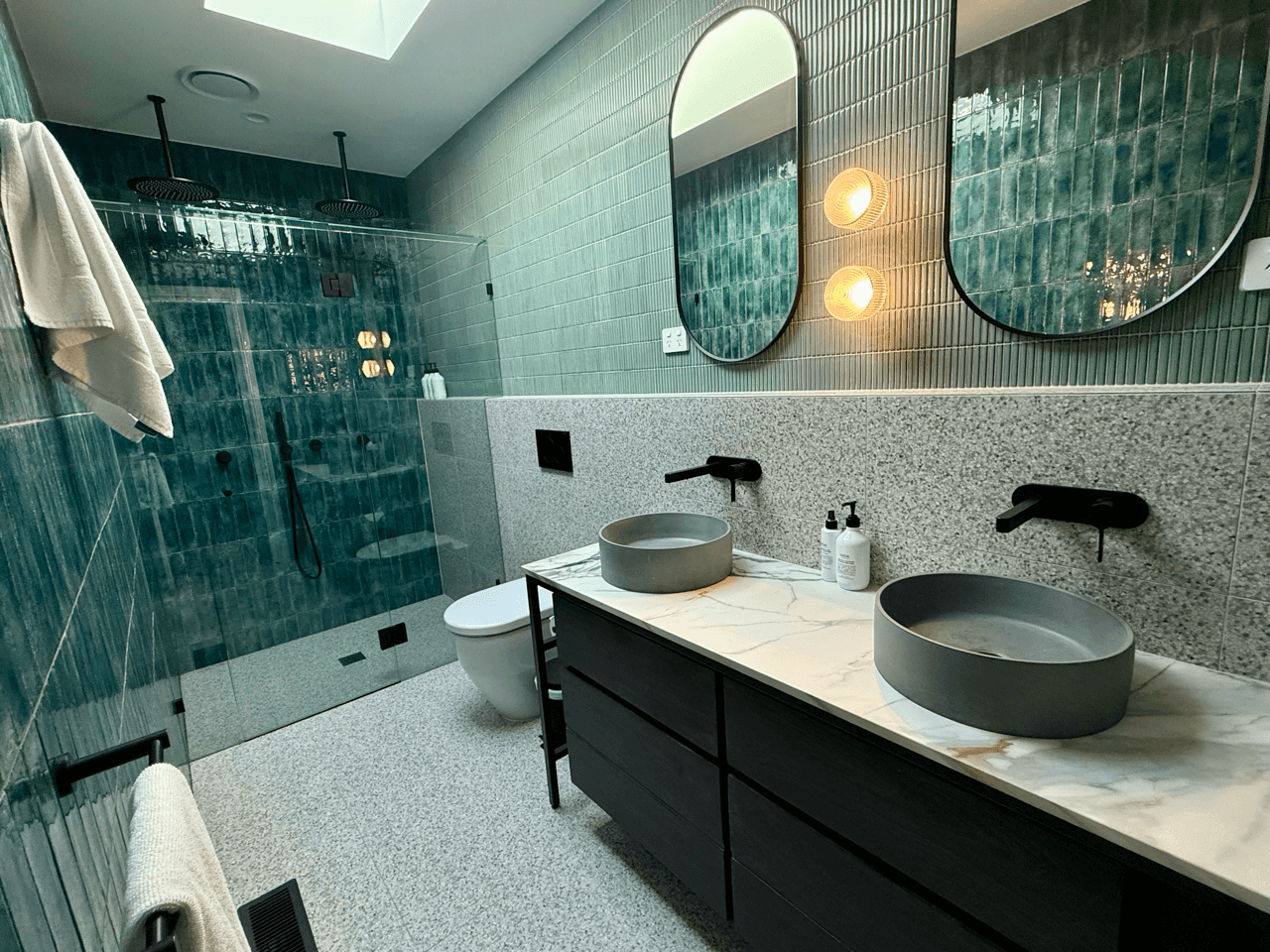Modern bathroom with green tiled walls, a double sink vanity with black cabinetry, circular gray vessel sinks, black wall-mounted faucets, a large mirror, a skylight, and a walk-in shower with black rain showerheads and a glass partition.