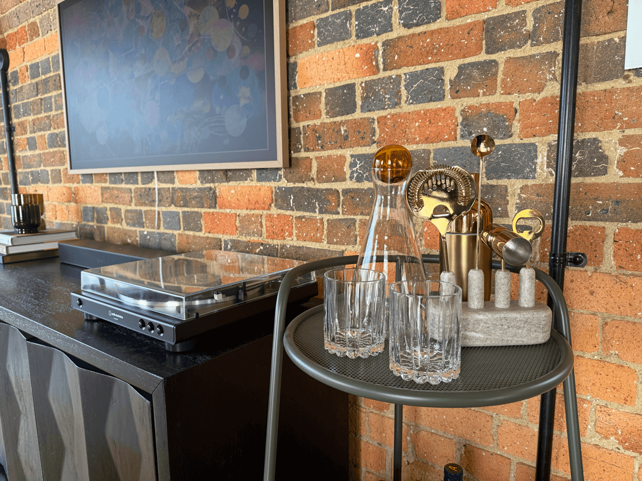 Record player and bar cart with four empty glasses, decanter, and cocktail stirrers in a holder, set against an exposed brick wall.
