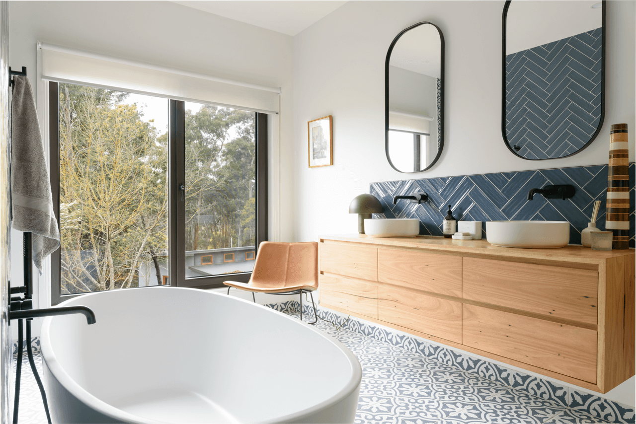 Modern bathroom with freestanding bathtub, wooden double vanity with vessel sinks, two mirrors, large window showing trees outside, patterned floor tiles, and a brown chair.