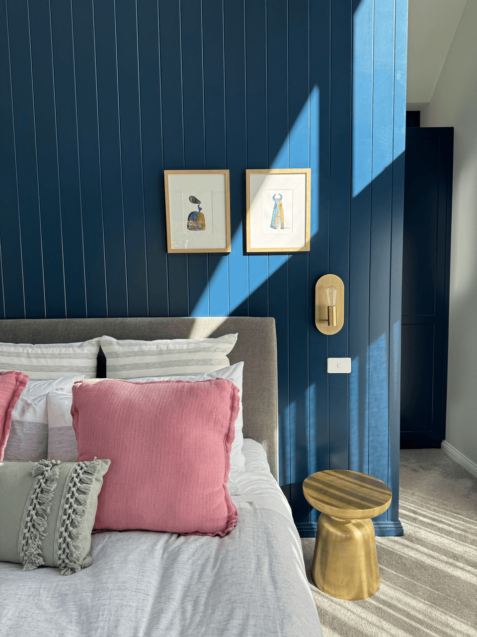 A bedroom with a blue paneled wall, framed artwork, a beige bed with pink and beige pillows, a gold side table, and sunshine creating shadows on the wall.