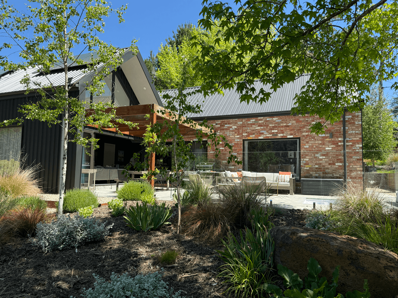 A backyard patio area with outdoor seating, surrounded by plants and trees, and a modern house with brick and metal exterior in the background.