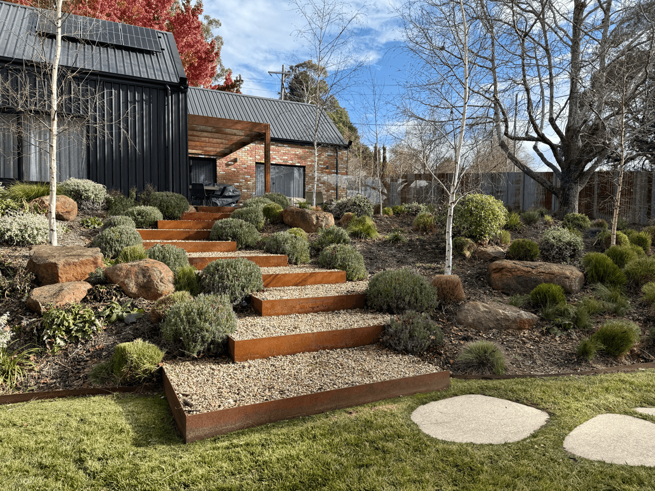 Backyard with metal stairs leading up a landscaped garden containing shrubs, rocks, and leafless trees, with a black and brick house in the background under a partly cloudy sky.