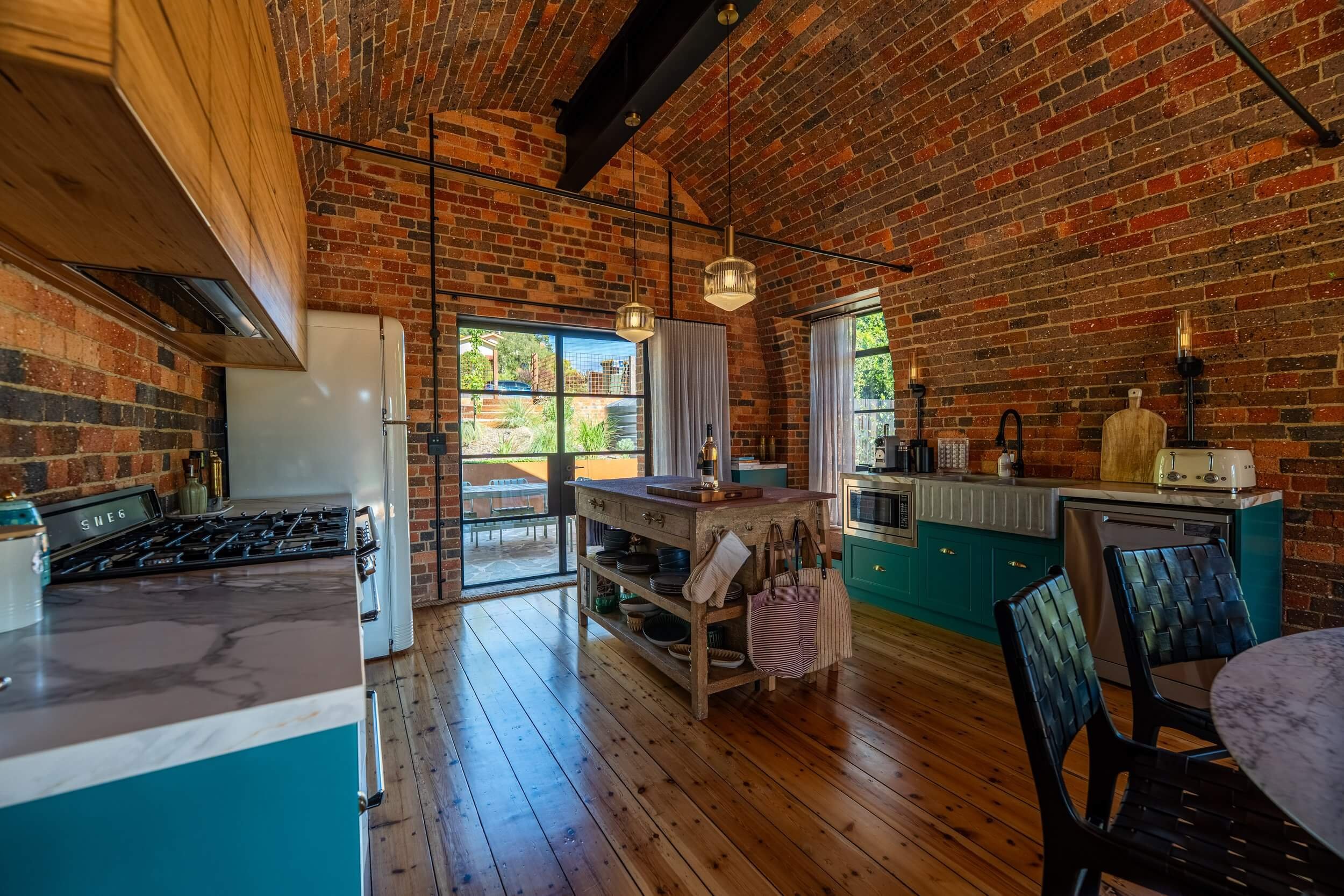 A kitchen with red brick walls and vaulted ceiling, wooden flooring, and large windows. Contains a wooden island, teal cabinets, vintage appliances, and a round marble table with black chairs. Hanging pendant lights provide lighting.