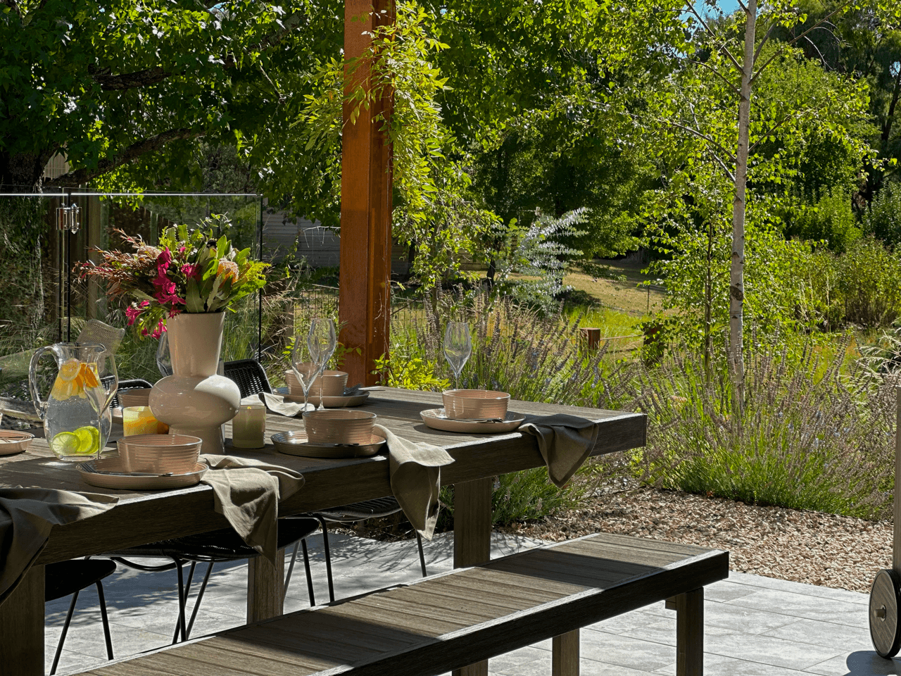 An outdoor dining table set for a meal, with a large white vase of pink flowers, glasses, bowls, and plates. The table is on a patio with a garden and trees in the background.