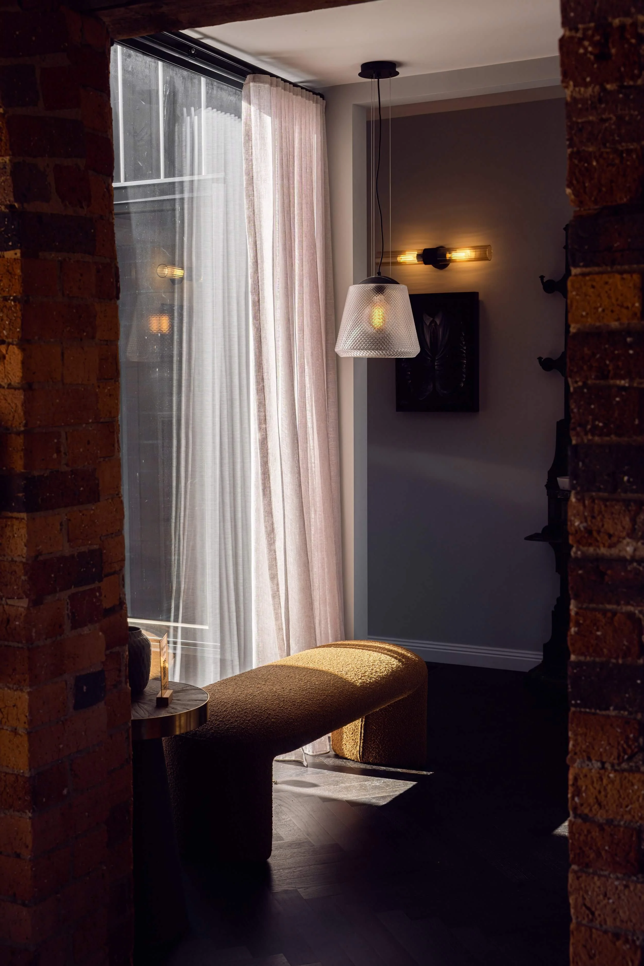 A cozy corner of a room featuring a window with sheer white curtains, a textured pendant light, a framed artwork on the wall, and a mustard-colored ottoman with a side table and decorative items, framed by exposed brick walls.