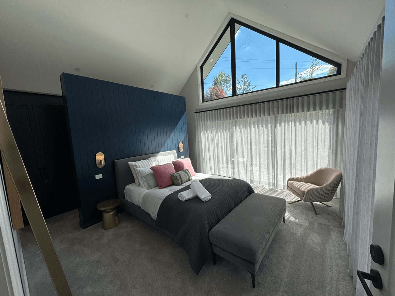 Modern bedroom with a large window, blue accent wall, bed with pink pillows, ottoman at the foot of the bed, and a beige armchair near the window.