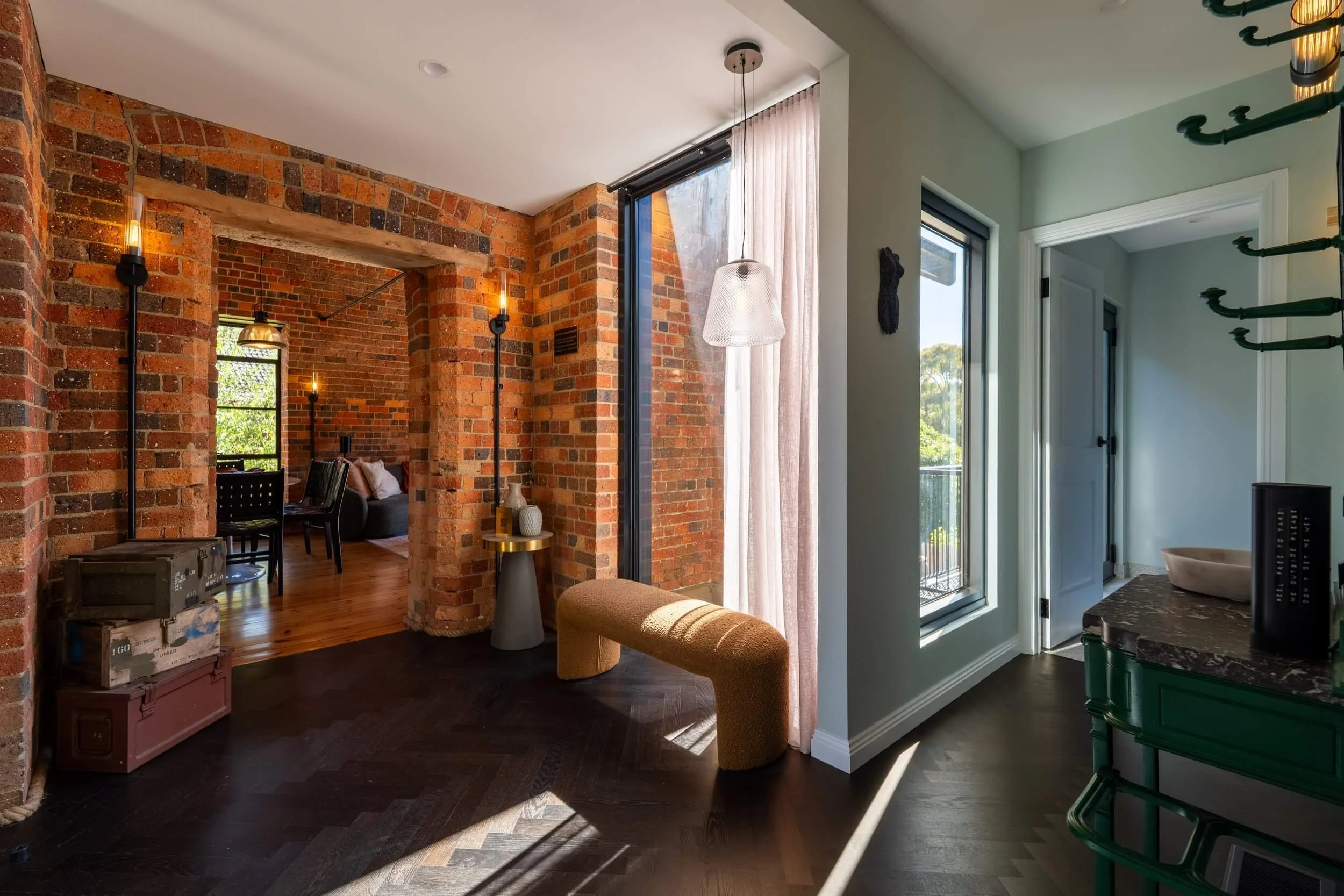 Interior view of a modern apartment with exposed brick walls, large windows with curtains, and contemporary furniture including a small table, a bench, and decorative items. Sunlight streams into the space, highlighting the dark hardwood floors.