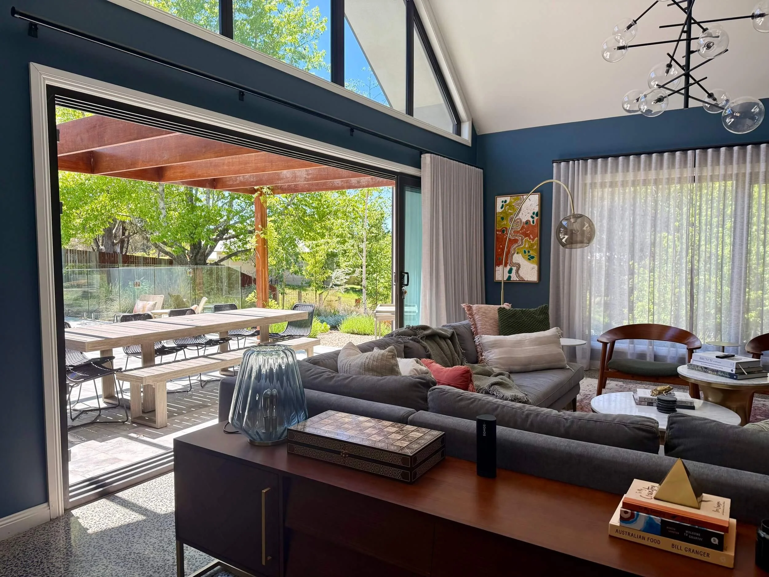 Living room with large sliding glass door leading to outdoor patio, which has a wooden dining table and chairs under a wooden pergola. Inside, there is a gray sofa with pillows, a small table with books and decor, a modern floor lamp, and curtains ov