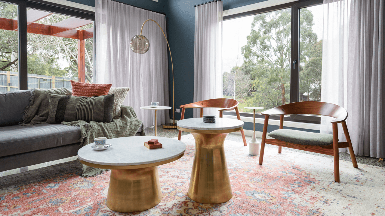 Modern living room with large windows, a gray sofa with pillows, two round tables with marble tops and gold bases, two wooden armchairs with green cushions, a standing floor lamp with a glass shade, and sheer white curtains, with a view of trees outs