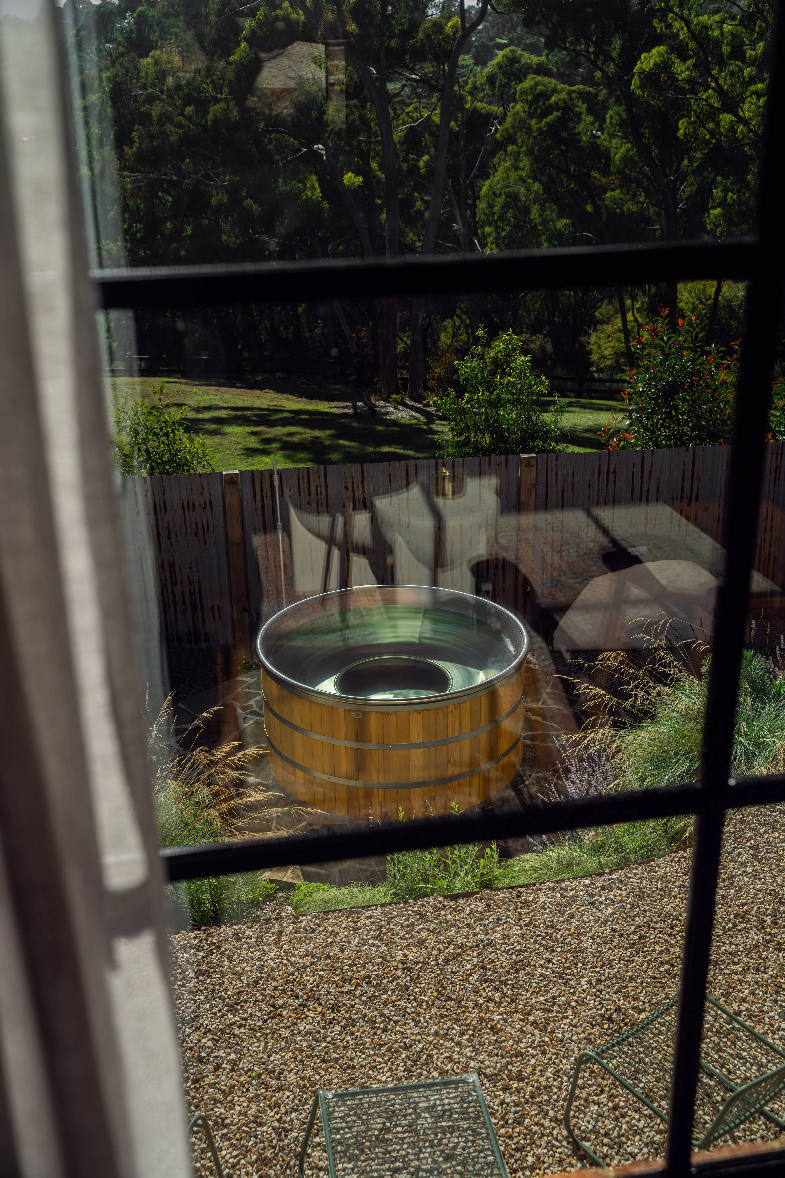View through a window showing a wooden hot tub outside on a gravel patio, surrounded by plants and trees, with a backyard fence and lush green trees in the background.