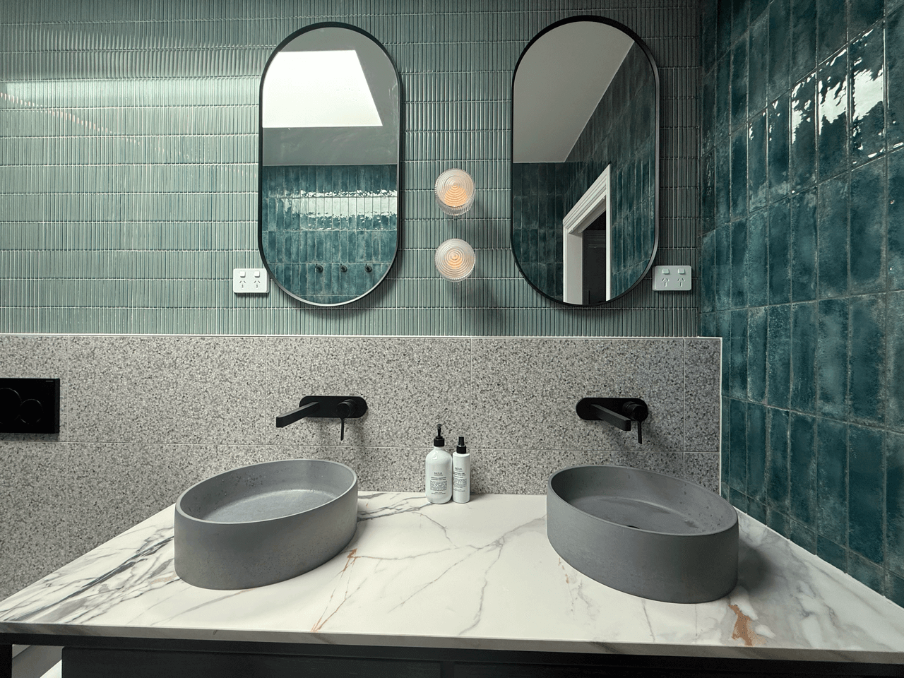 Modern bathroom with dual grey vessel sinks on a white marble countertop, green tiled wall with two oval mirrors, black faucets, and two bottles of soap or lotion on the counter.