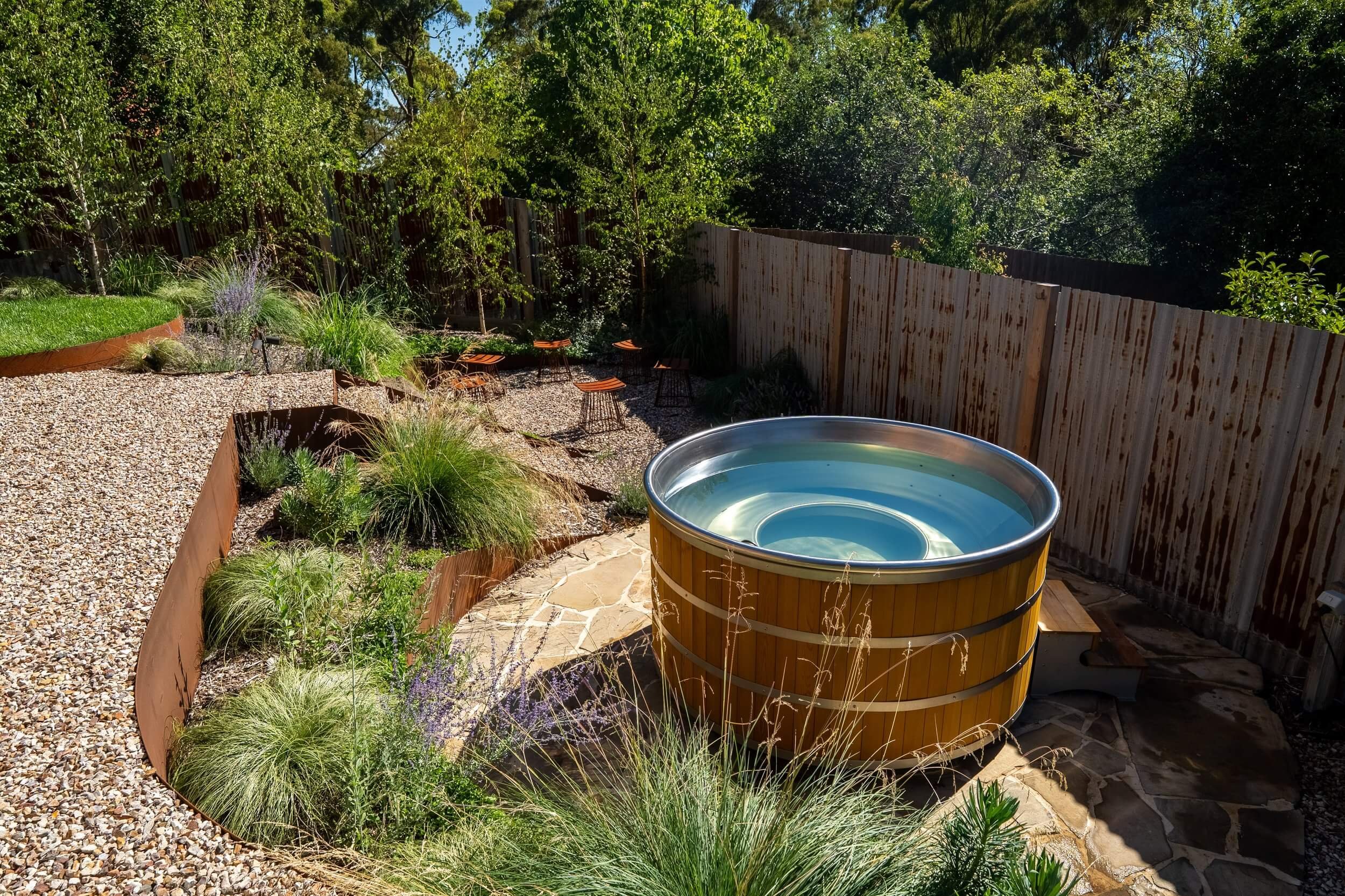 A backyard garden with a water barrel, gravel pathway, and wooden fence surrounded by green trees and plants.