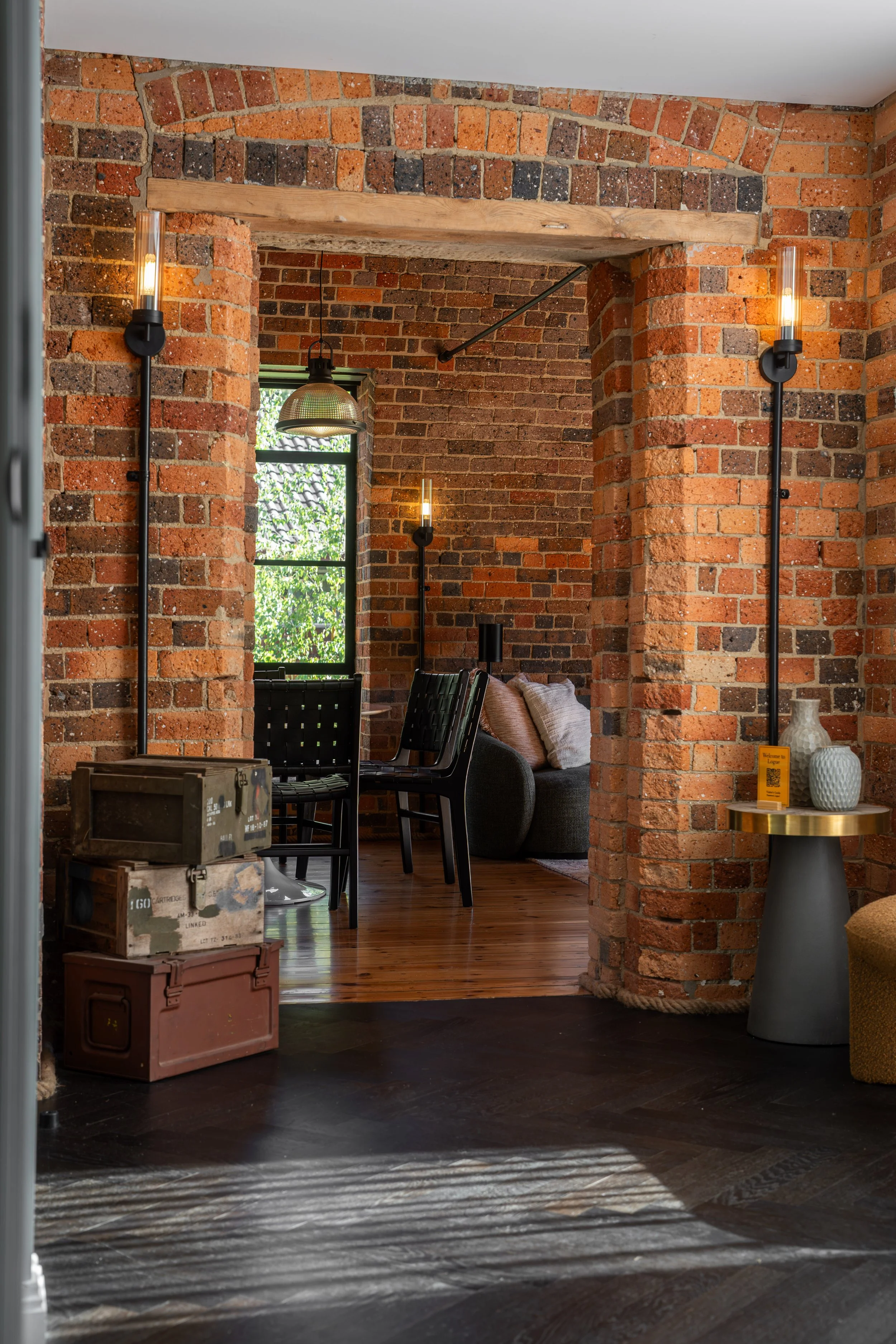 Interior view of a room with brick walls, wooden beams, and contemporary lighting fixtures, including wall-mounted lights and an overhead industrial-style pendant light, with a seating area featuring a couch and chairs, and decorative vases on a smal