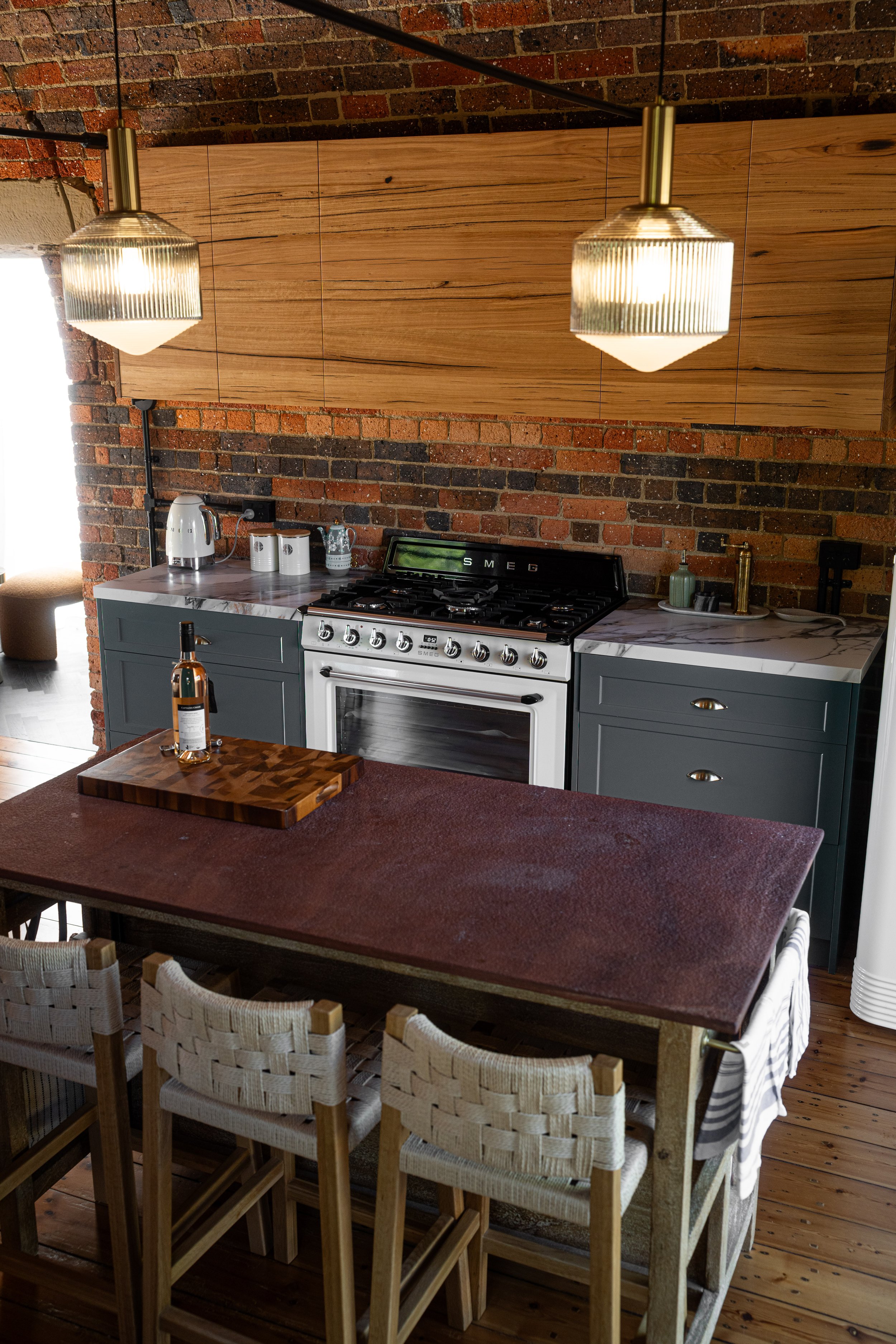 Modern kitchen with brick wall, dark green cabinets, white stove, marble countertops, and wooden ceiling. There are two hanging pendant lights, a wooden table with chairs, and various small appliances on the counter.