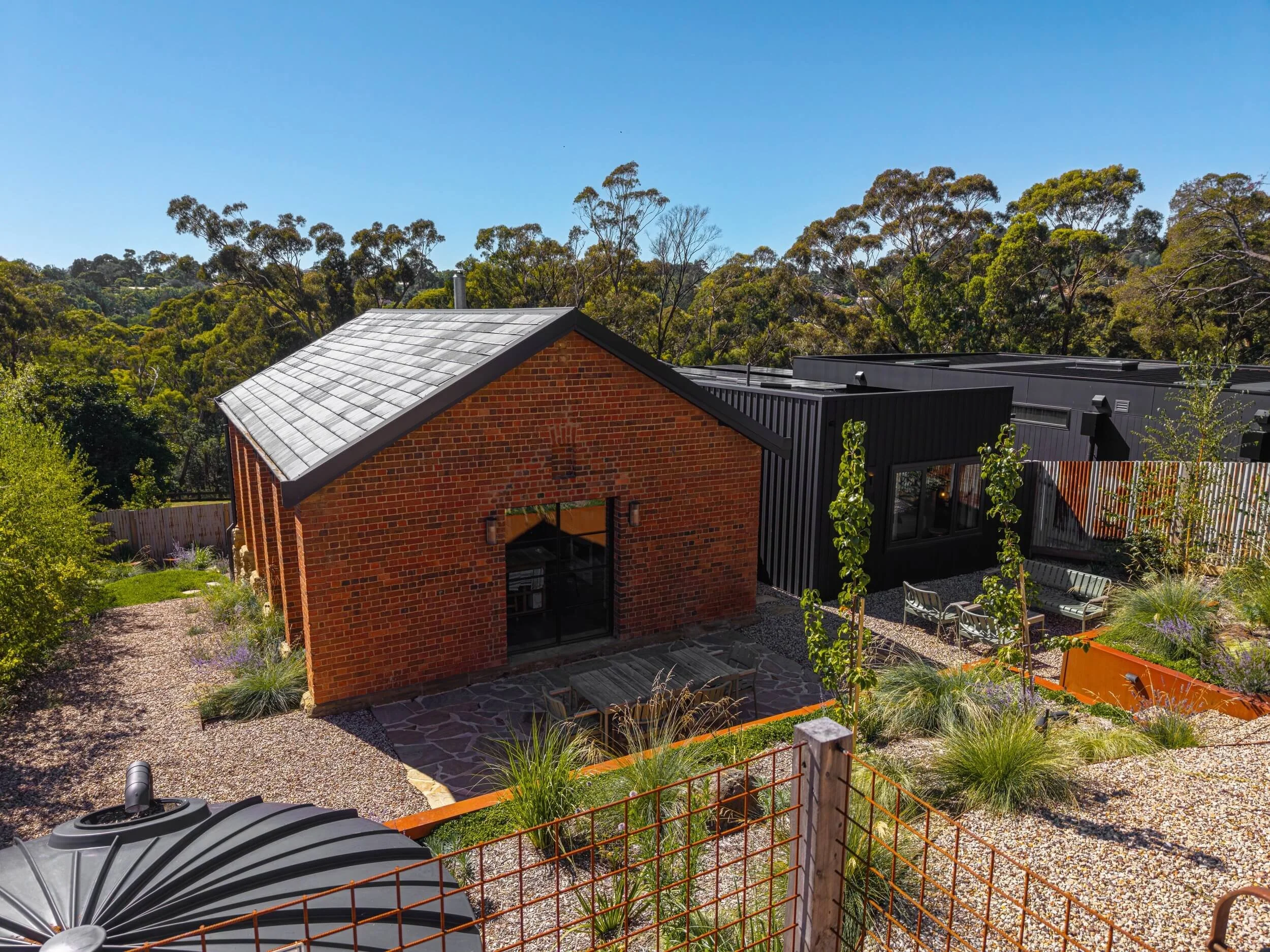 A modern house with a brick wall and black roof, surrounded by a garden with trees, plants, and gravel paths under clear blue sky.