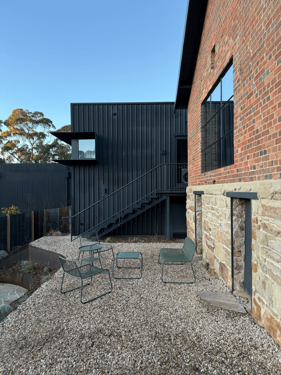 Modern house exterior with a black metal and brick facade, outdoor seating area with four chairs and a small table on gravel ground, staircase leading to an upper level, trees in the background, and a clear blue sky.
