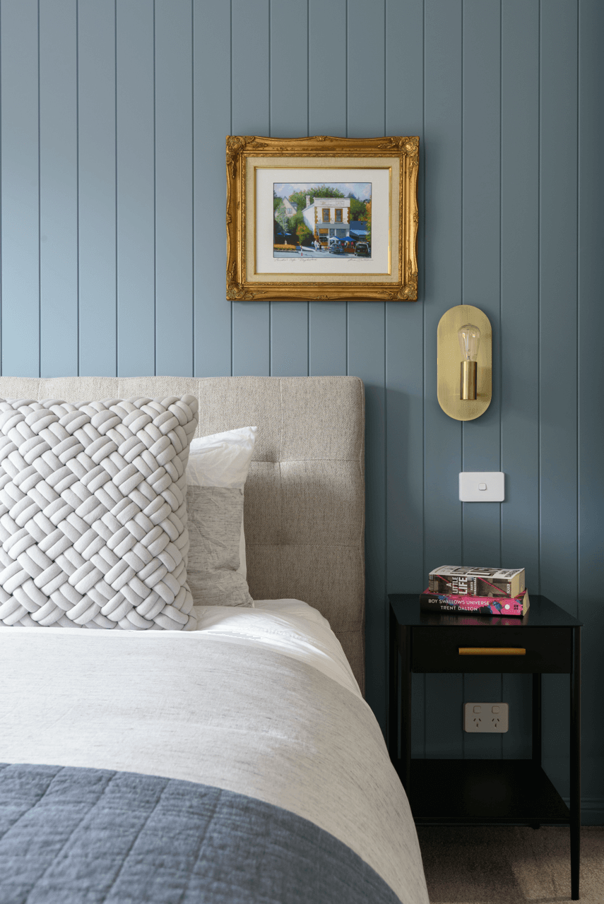 A cozy bedroom corner with a bed featuring a textured white pillow and beige headboard, a black nightstand with books, a blue paneled wall decorated with a framed painting of a building and a wall-mounted light fixture with a visible light bulb.