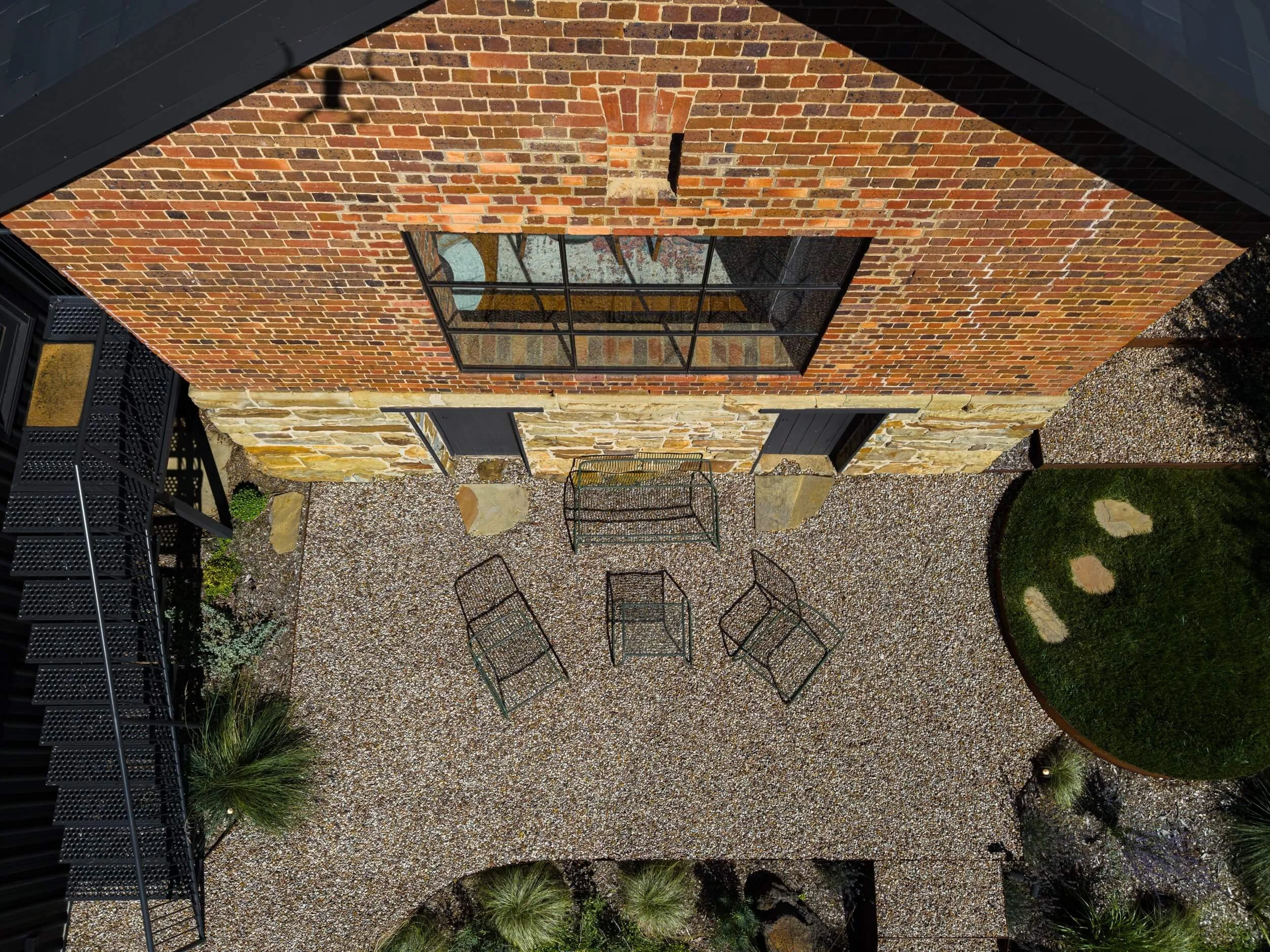 A backyard patio view from above, showing a brick house wall with a large window, outdoor chairs, gravel ground, and landscaping with grass and plants.