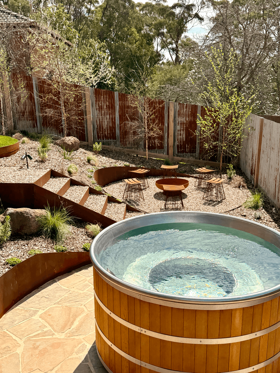 View of a backyard with a hot tub in the foreground, gravel seating area with chairs and tables, surrounded by plants and trees, enclosed by a rusted metal fence.
