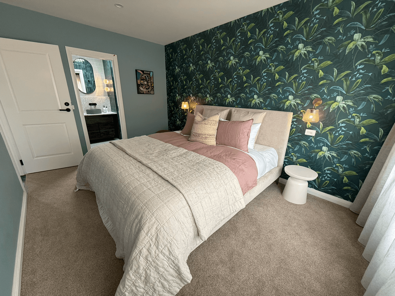 A cozy bedroom with a large bed featuring a beige upholstered headboard, decorated with pink, beige, and white pillows, a beige quilted blanket, and a pink bed runner. The wall behind the bed has dark green leafy patterned wallpaper, and there are wa