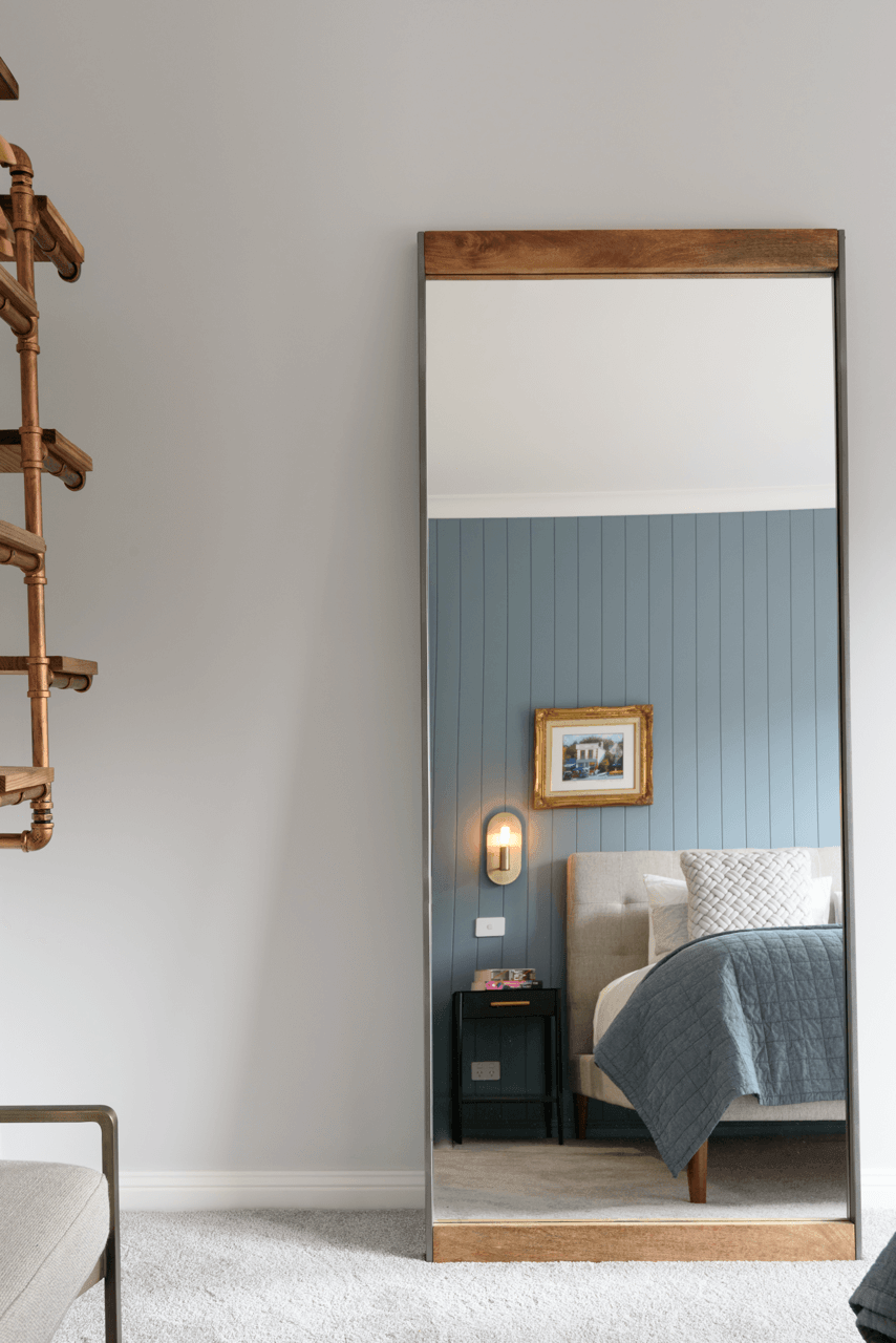 A full-length mirror reflecting a bedroom scene with a bed, nightstand, framed picture, and wall-mounted light fixture.