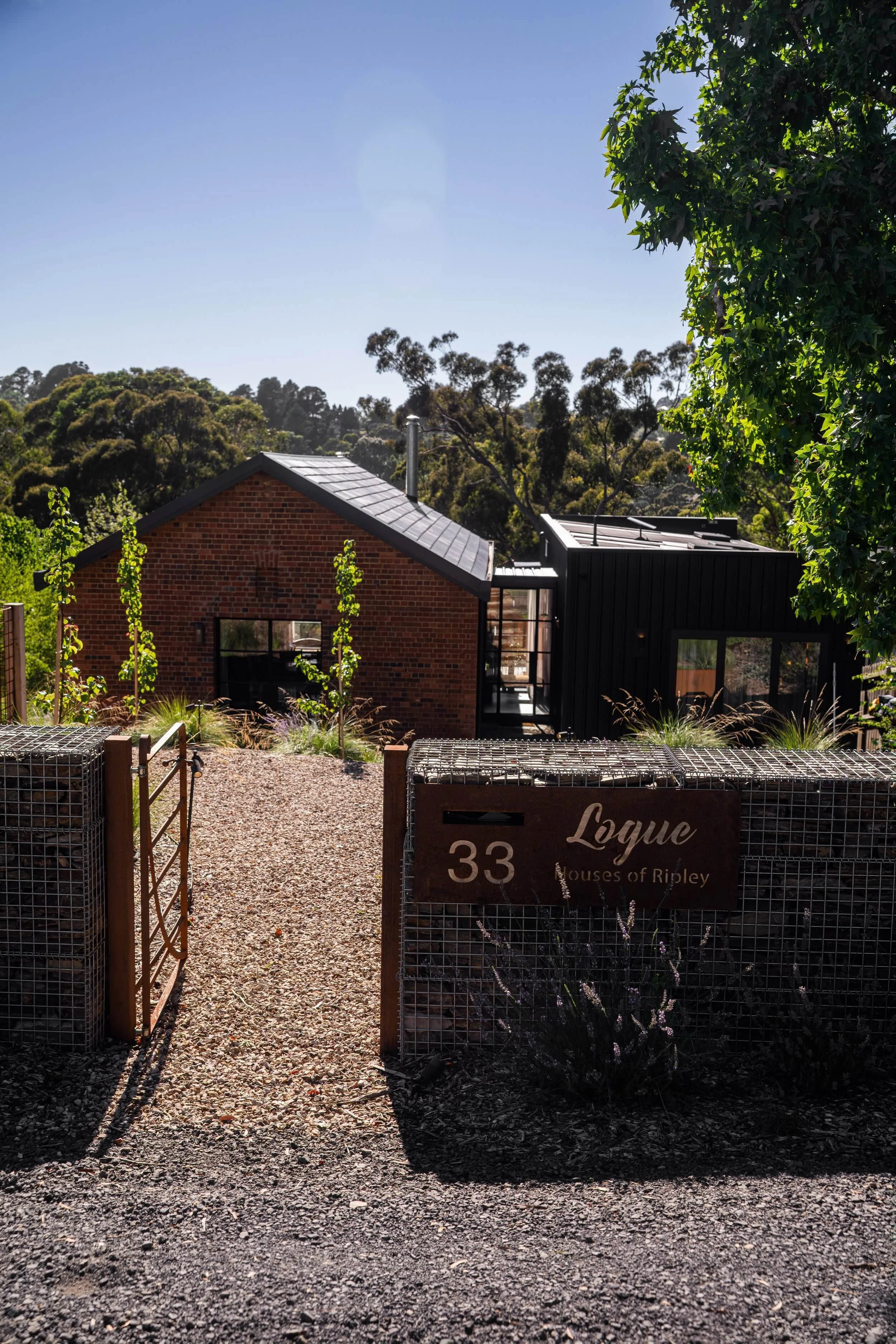 A modern house with a brick facade on the left and black siding on the right, surrounded by a gravel pathway and a garden with small trees and plants. There is a wooden gate with a sign that reads 'Loyue, Houses of Ripley' and the number 33. In the b