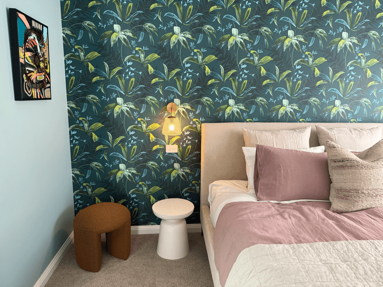 A bedroom with a bed, pillows, and a dark green leafy patterned wallpaper. There is a wall-mounted lamp beside the bed, a small brown stool, and a white round side table. A framed colorful art piece hangs on the light blue wall.