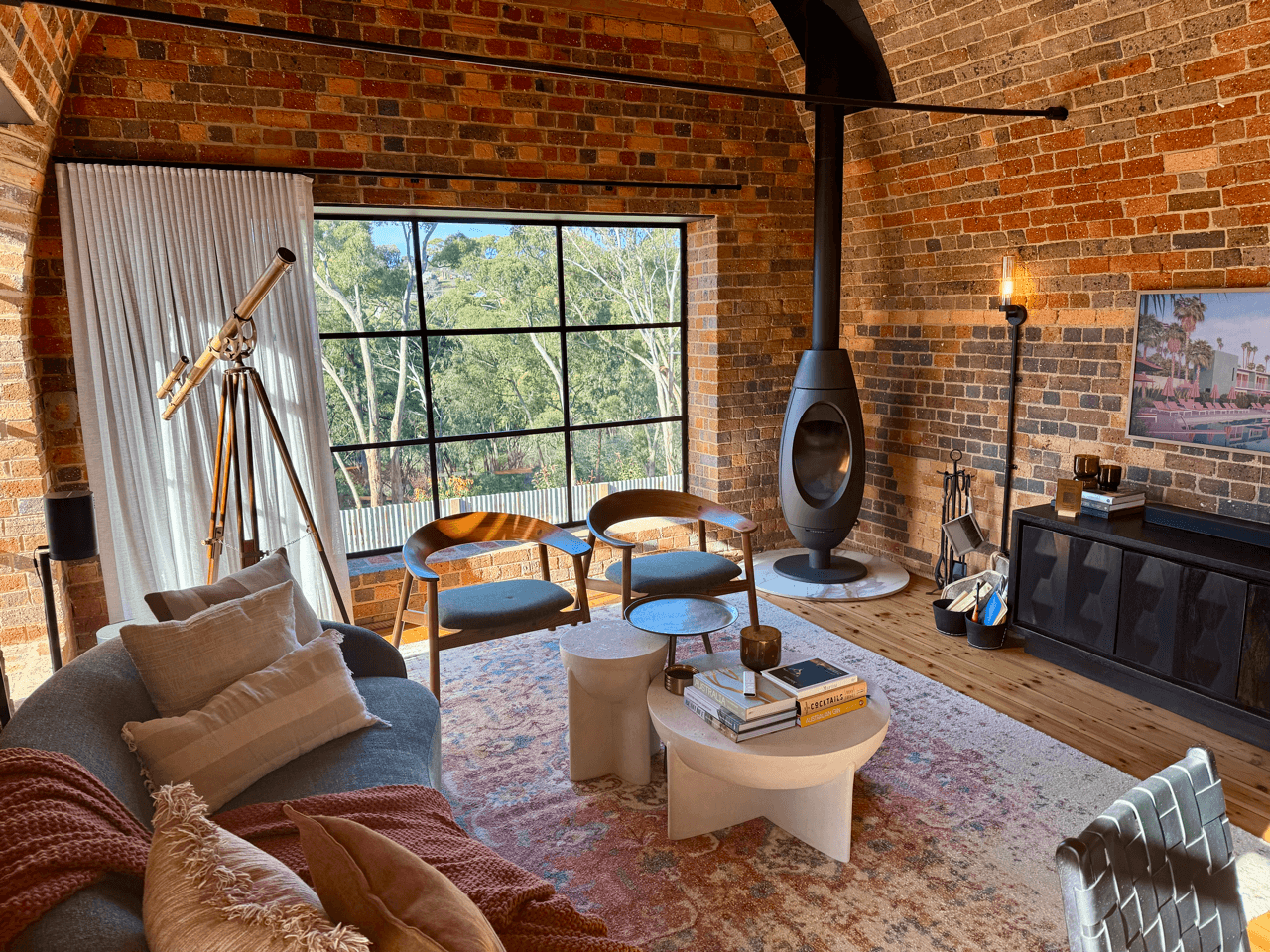 Living room with brick walls, large window, modern fireplace, white coffee table with books, gray sofa with pillows, artwork on wall, wooden floor, and telescope near window.