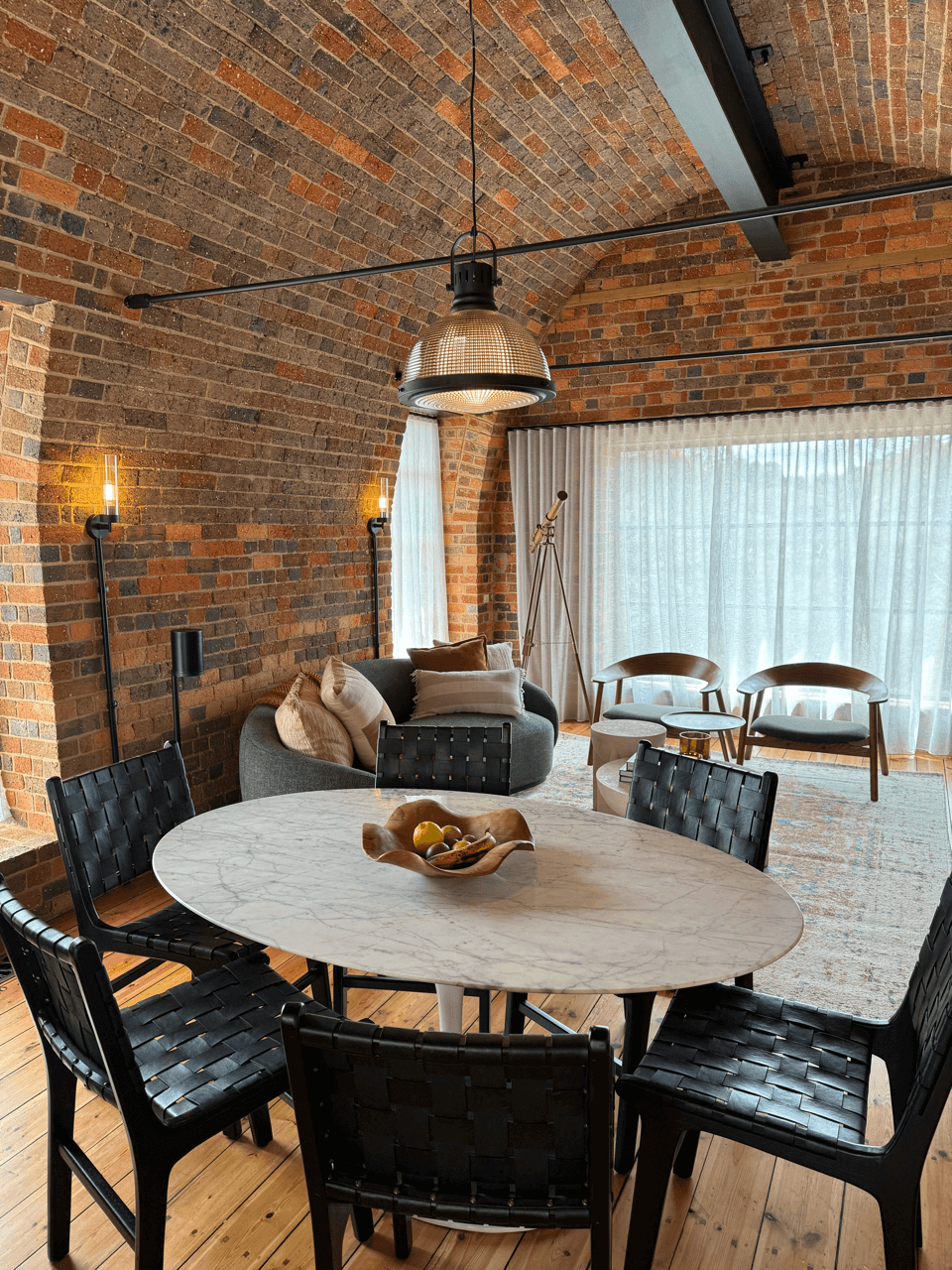 Modern living room with brick vaulted ceiling, round marble dining table surrounded by six black woven chairs, gray sofa with pillows, telescope near large window with white curtains, wooden chairs and small table, and contemporary lighting fixtures.