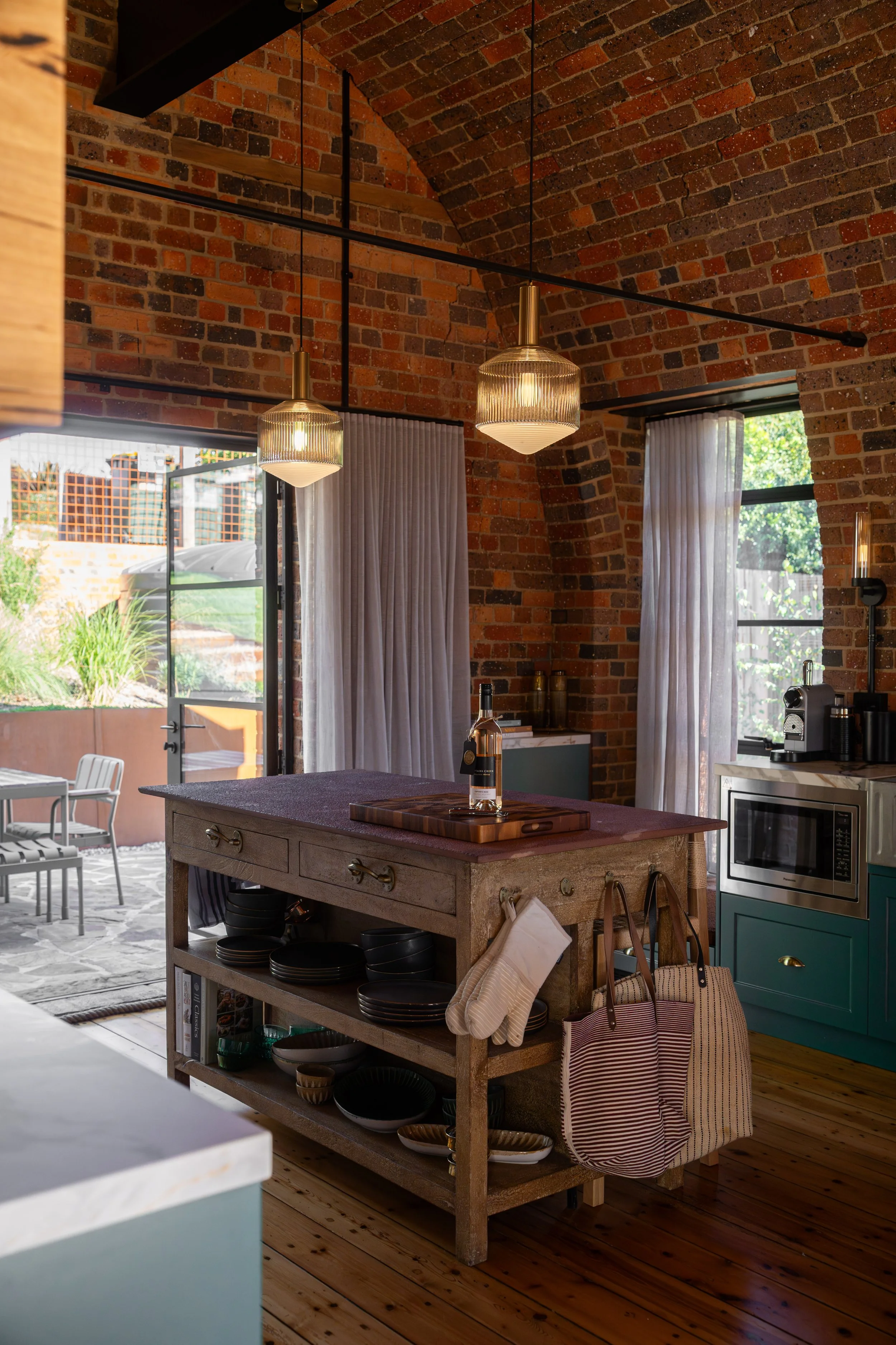 A rustic kitchen with exposed brick walls, pendant lighting, and a kitchen island with plates underneath. An open door leads to an outdoor patio with seating and greenery. Kitchen appliances and a window with sheer curtains are visible.