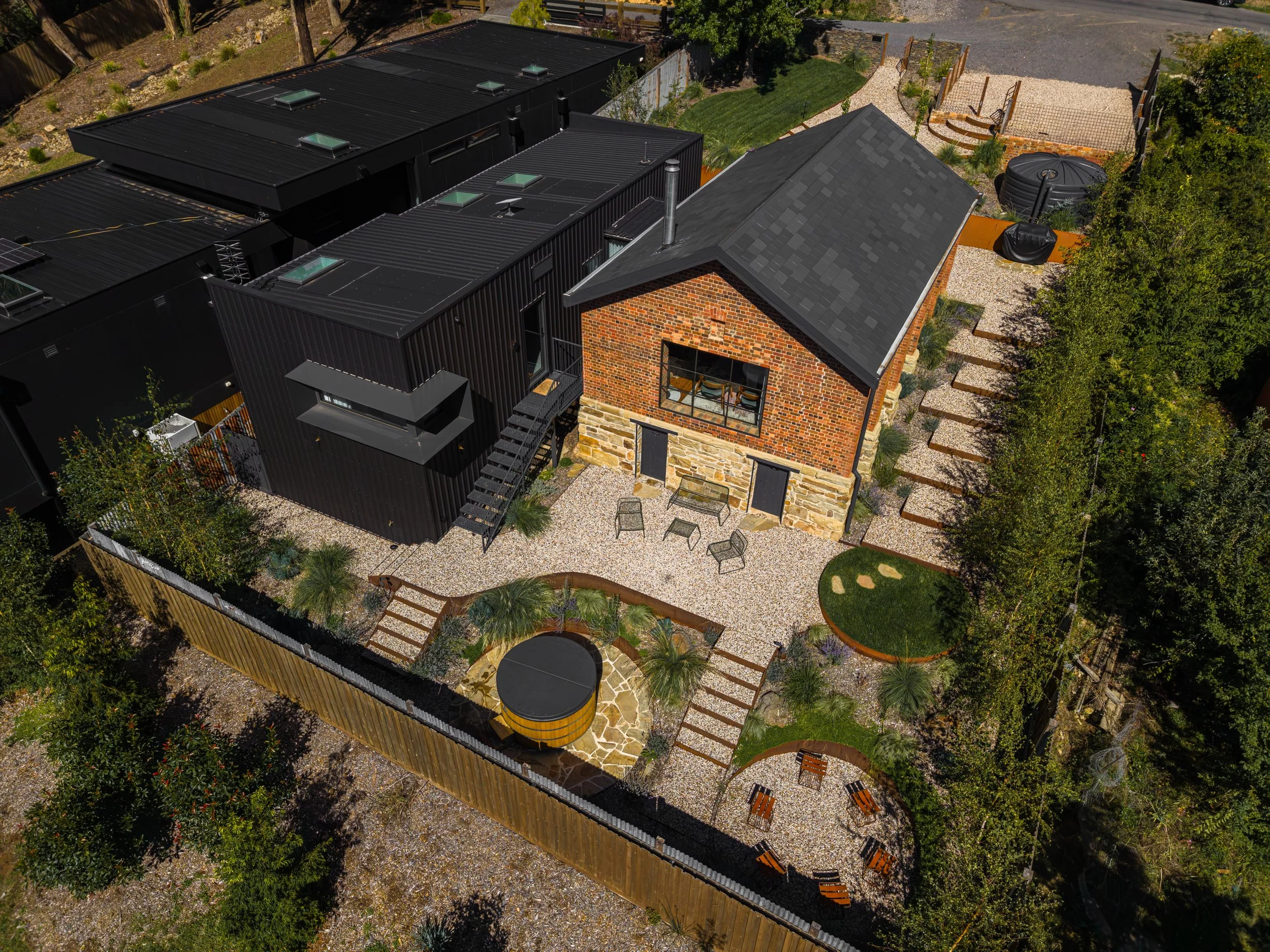 Aerial view of a suburban backyard featuring a historic brick and stone house with a slate roof, a patio with outdoor furniture, a small circular grassy area, a hot tub, garden beds, trees, and pathways, enclosed by a wooden fence.