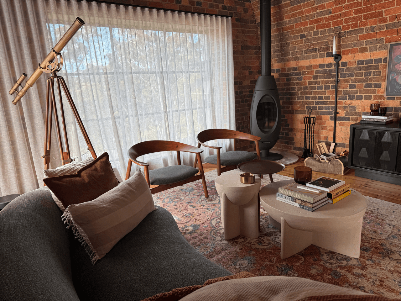 Living room with floor-to-ceiling sheer curtains, two wooden armchairs with gray cushions, a gray sofa with throw pillows, a modern stone coffee table with books, a small round side table with a small decorative item, a fireplace with accessories, a 