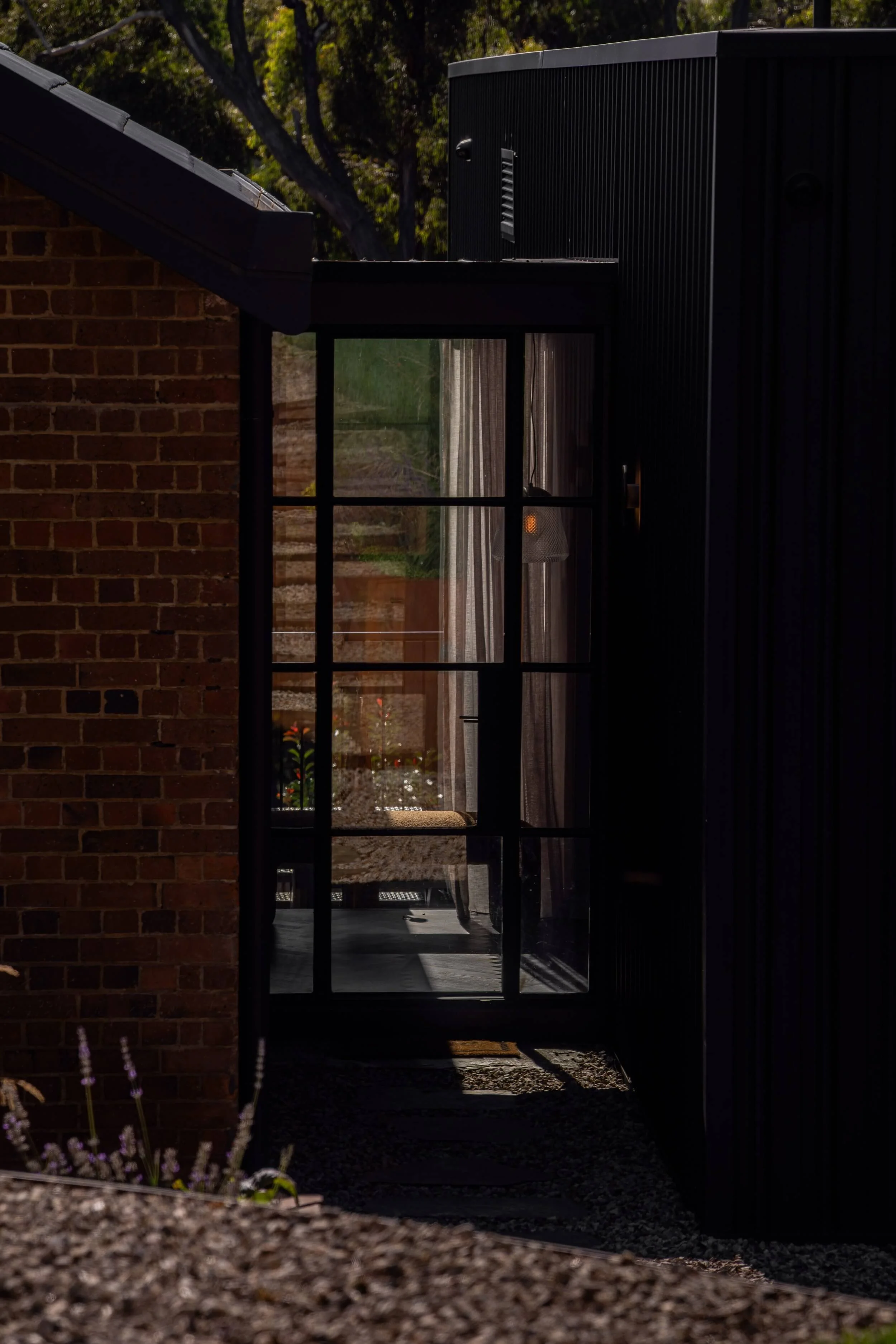 A glass door leading into a room with a view of a backyard garden with trees, plants, and flowers, through a large window. The door is open, showing a patio area outside. Inside, there is a hanging light fixture and curtains.