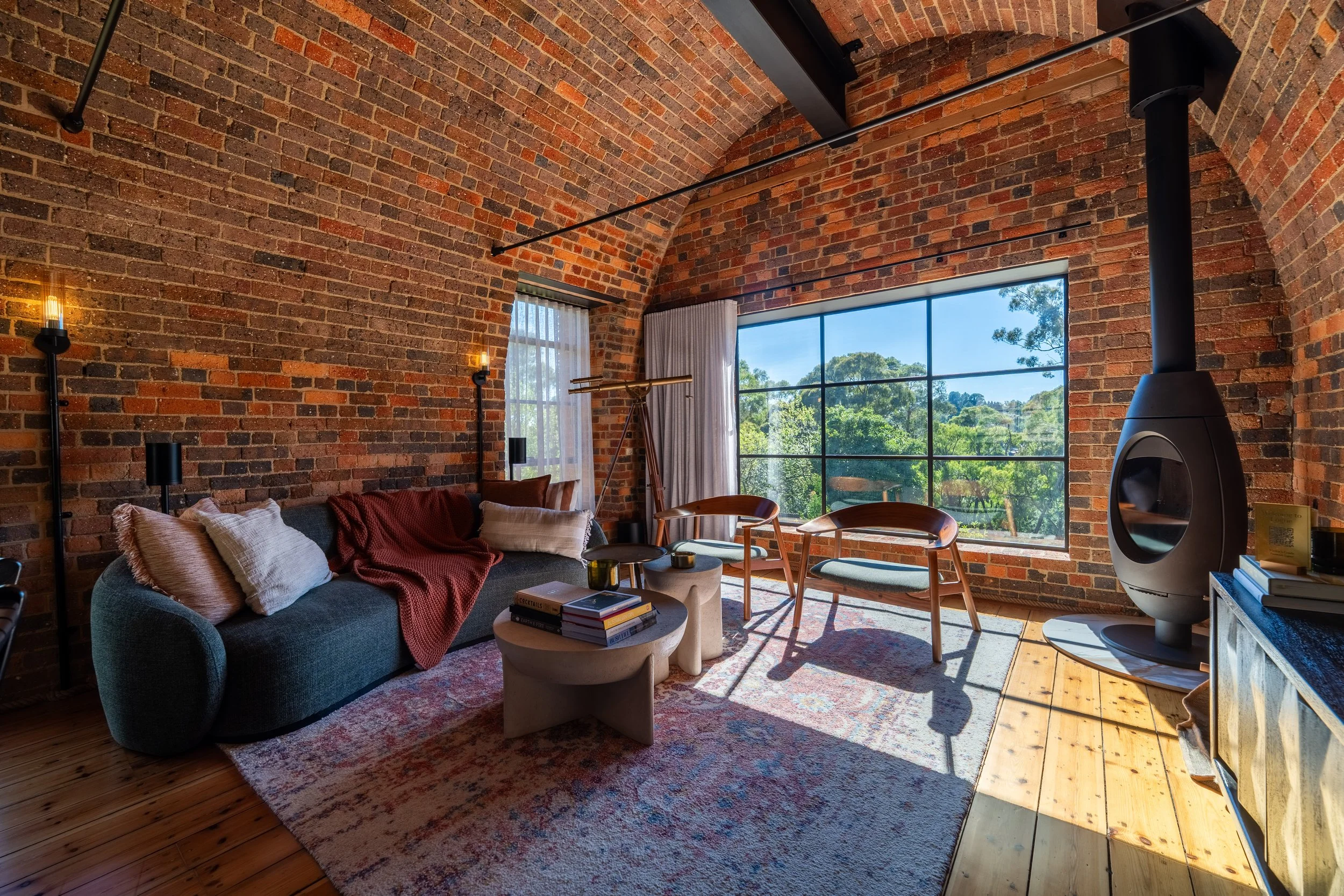 Living room with exposed brick domed walls, large window showing trees outside, modern furniture including a gray sofa with pillows and blanket, two wooden chairs, a telescope, and a black fireplace.