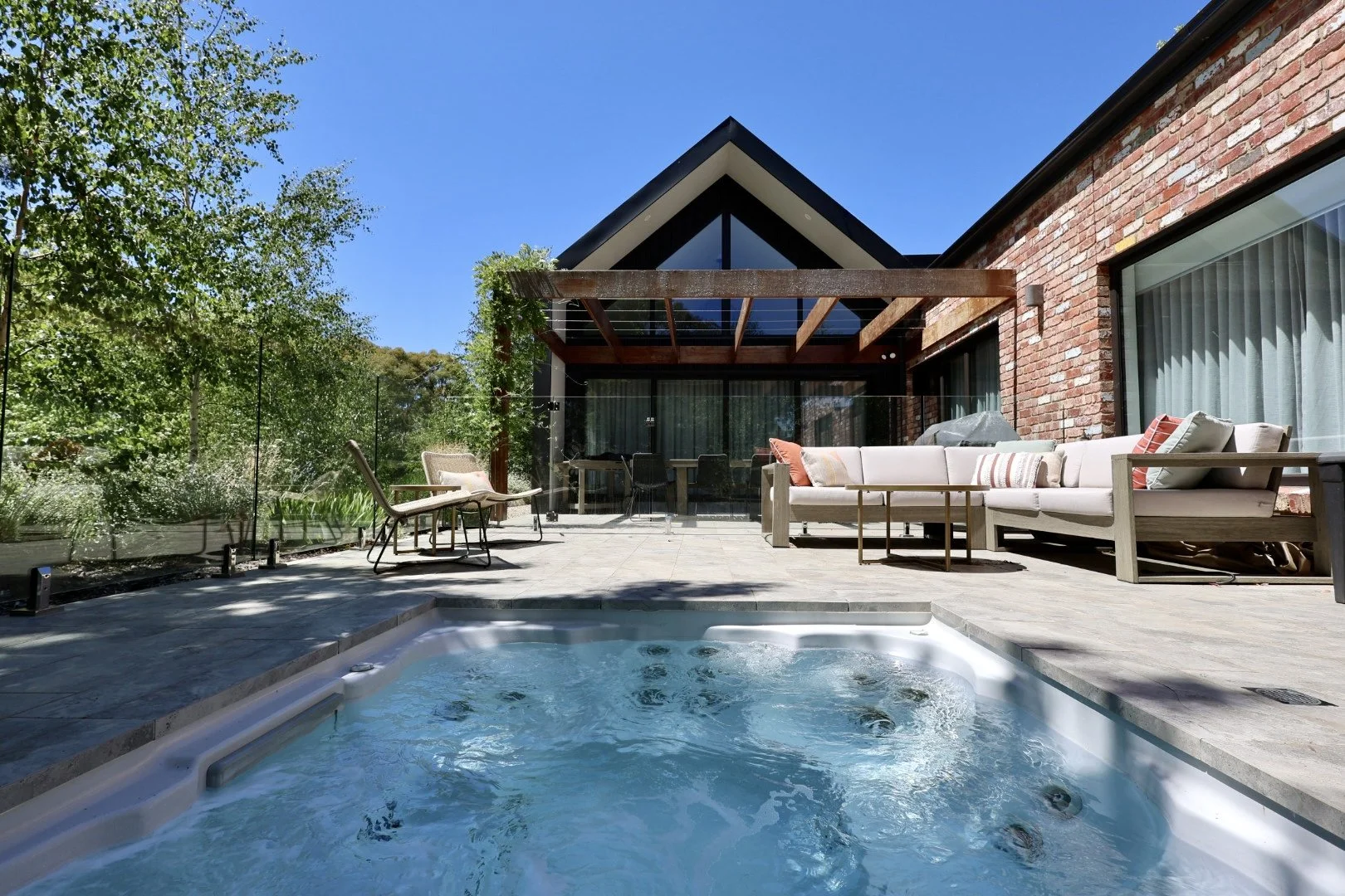 Backyard with hot tub, outdoor seating, brick house, trees, and clear blue sky.