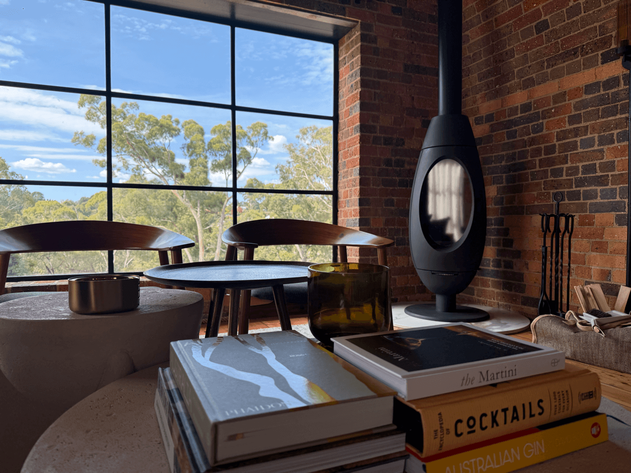 Interior of a modern room with a large window showing trees outside, featuring a stack of books on a table, a glass, a bowl, and a fireplace in the corner.