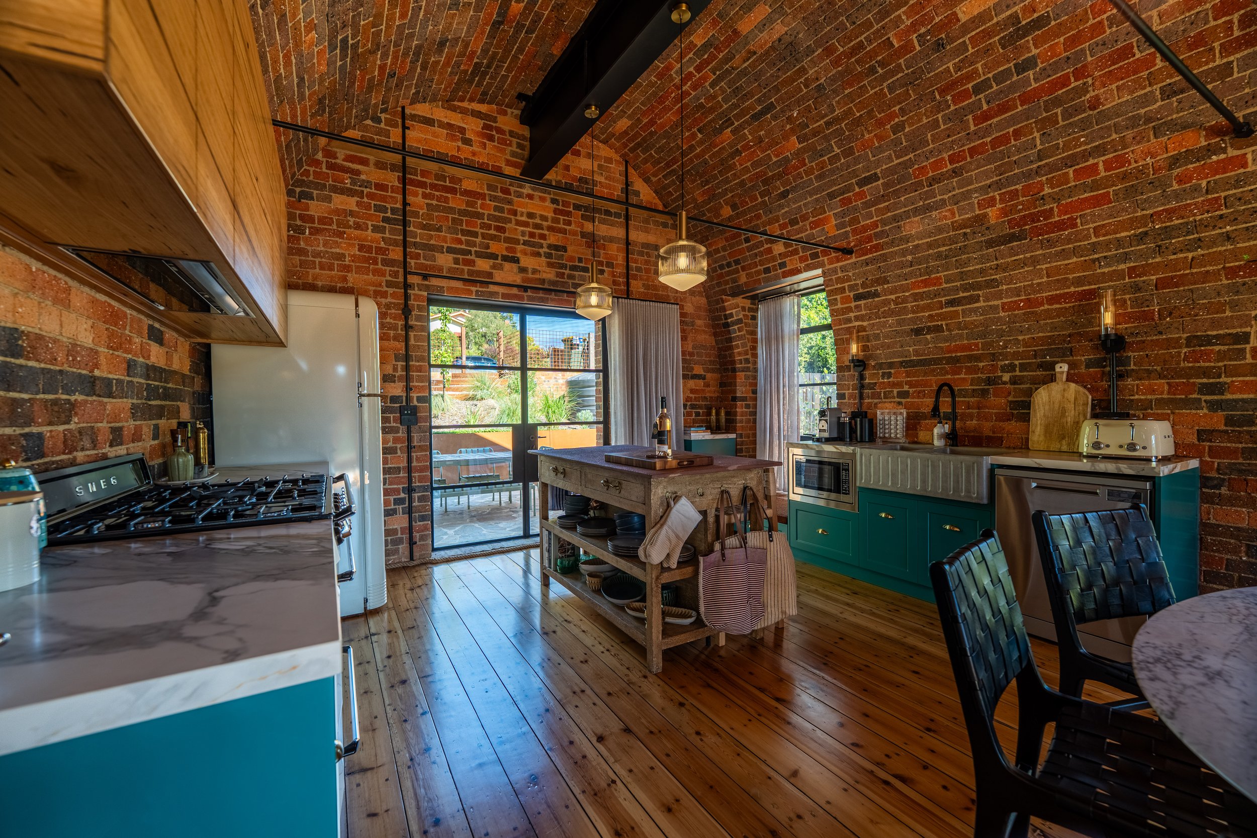 Kitchen with exposed brick walls, wooden floors, teal lower cabinets, white farmhouse sink, stove, refrigerator, island with open shelves, and a sliding glass door leading to an outdoor patio