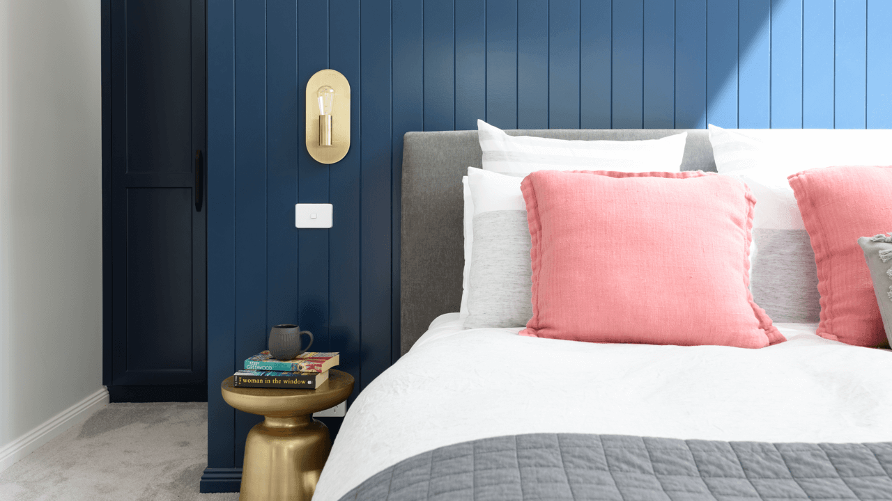 Close-up of a modern bedroom with a gray upholstered bed, pink and white pillows, a dark blue wall panel behind the bed, a small round gold side table with a stack of books and a black mug, a gold wall light fixture, and a light switch.