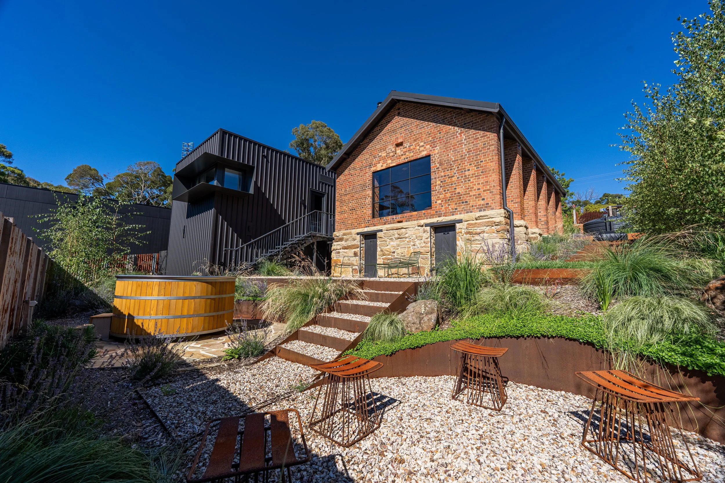 The image shows a backyard with a historic brick house and a modern black building. There are outdoor chairs, a hot tub, and a stone and gravel pathway surrounded by plants and landscaping.