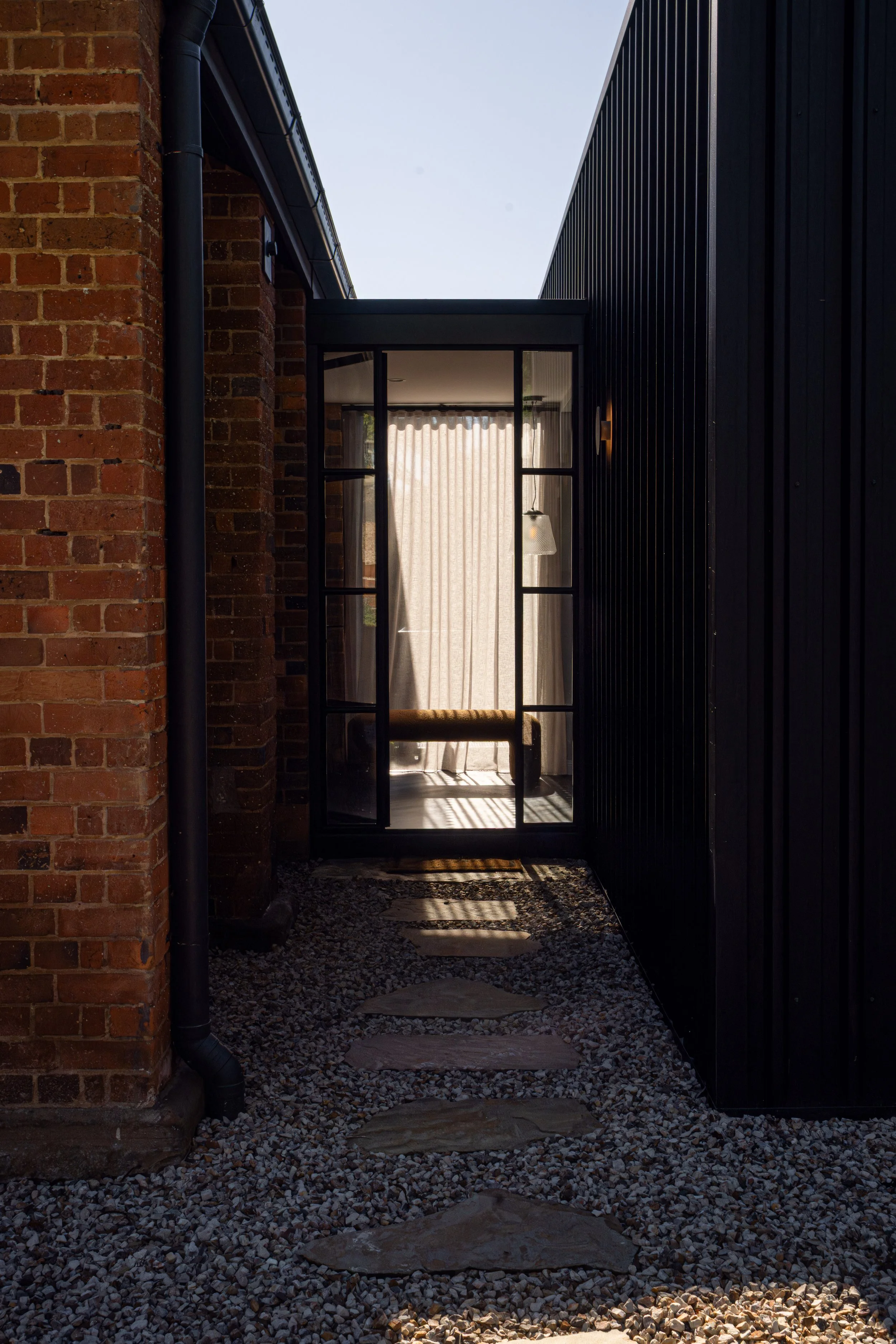 A narrow pathway with stone stepping stones leads to a glass door with black framing, revealing an interior with sheer curtains and a hanging light fixture. The pathway is flanked by a brick wall on the left and a black wooden wall on the right.