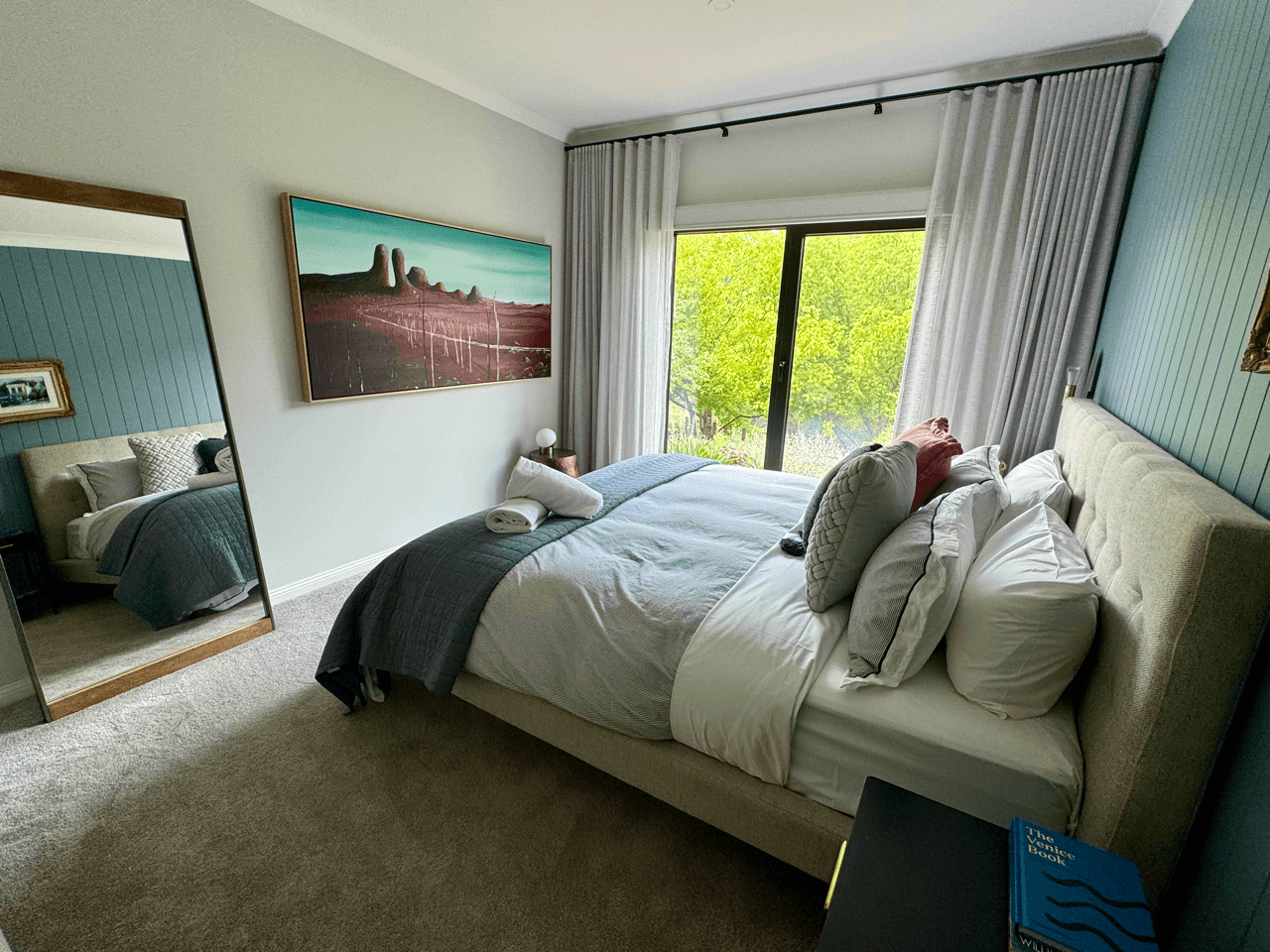 A neatly made bed with multiple pillows and a blanket in a bedroom with large window and curtains, a large mirror, a painting on the wall, and a nightstand with a book.